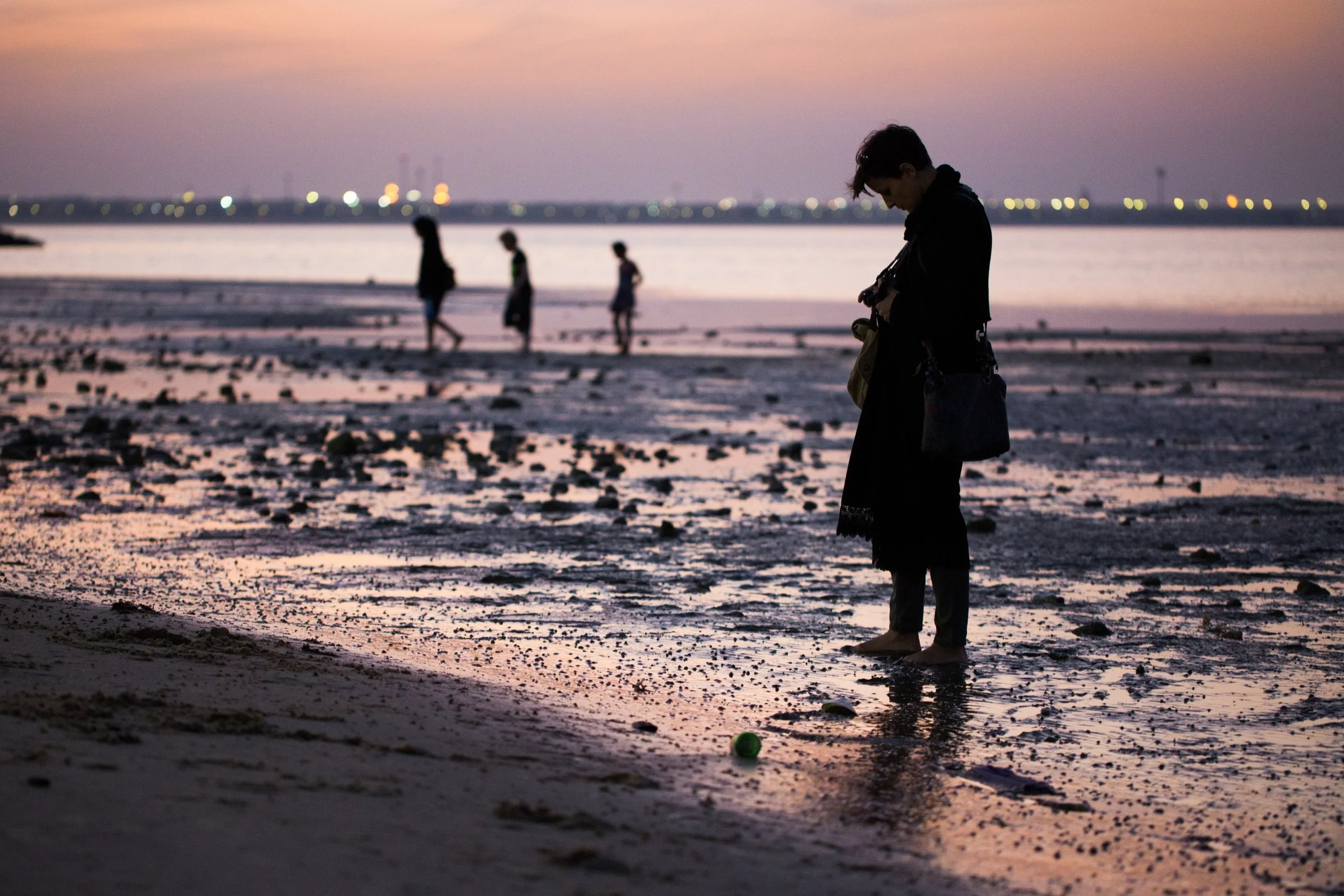 People walking along a beach at sunset, with one person in the foreground looking at a camera, and others in the background walking near the water. The sky has shades of pink and purple, and the beach has rocks and patches of water.