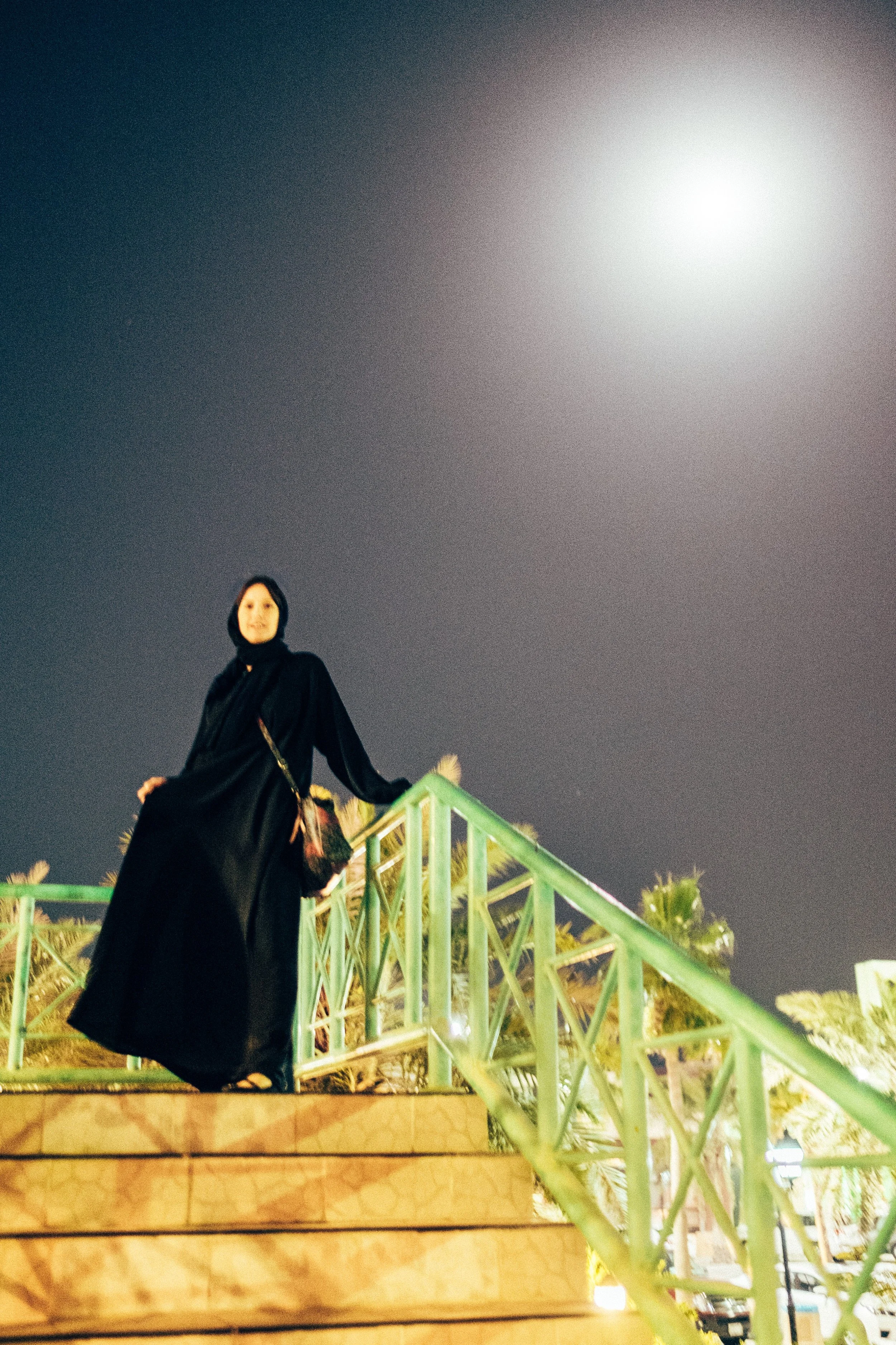 A woman dressed in black standing on outdoors illuminated steps at night, with a bright moon in the cloudy sky above. Palm trees and a green railing are visible around her.