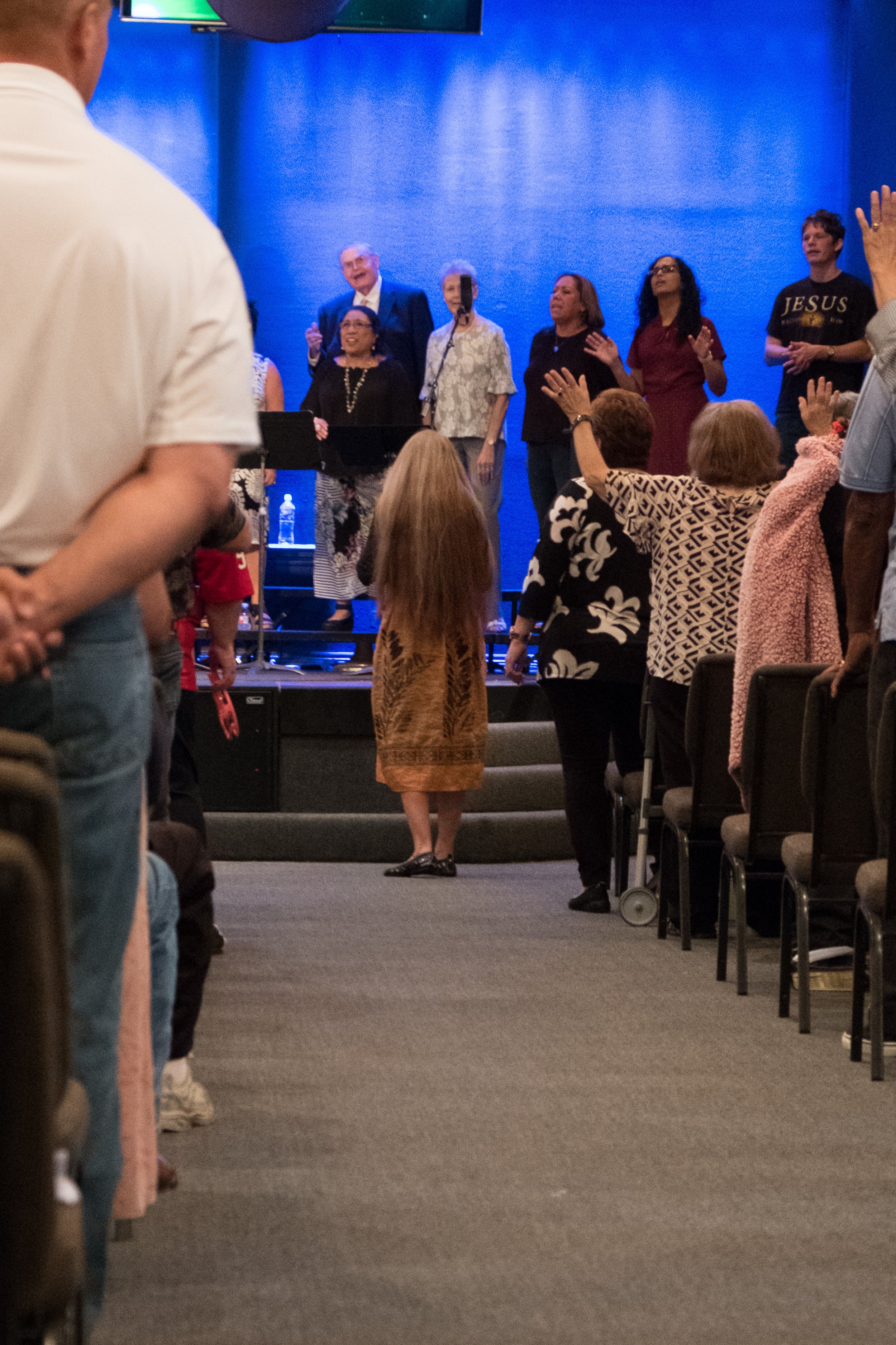 People gathered in a church or religious setting, with a woman at the front and a man near her on stage, all facing the congregation, and some congregation members raising their hands in worship or prayer.