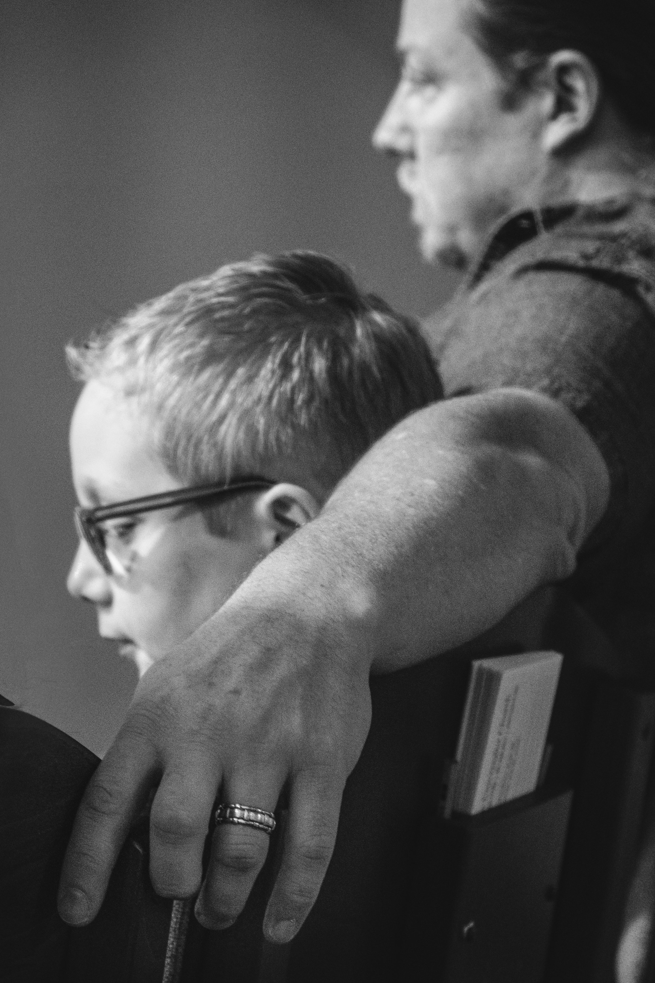 Close-up black and white photo of a person's hand resting on a bookshelf, wearing rings. Another person is in the background, partially visible, with long hair and a shirt, looking downward.