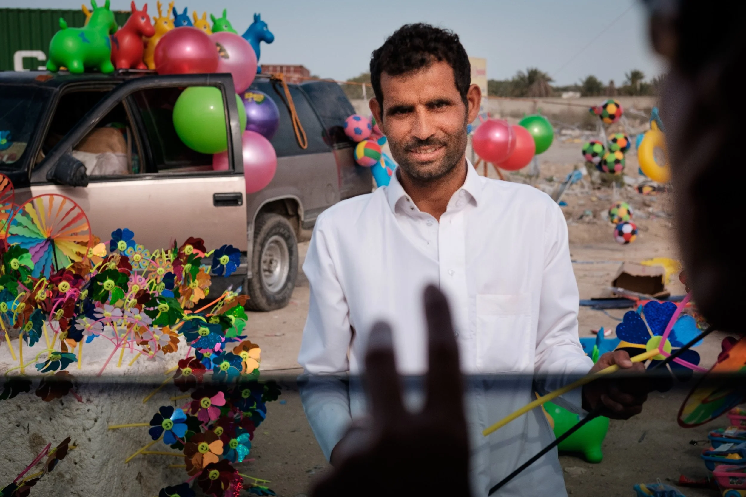 A man in a white shirt standing at a colorful outdoor market, with various toys and balloons, including green, pink, and purple balloons, around him. There is a car in the background decorated with inflatable toys like animals and balls, set in a dry