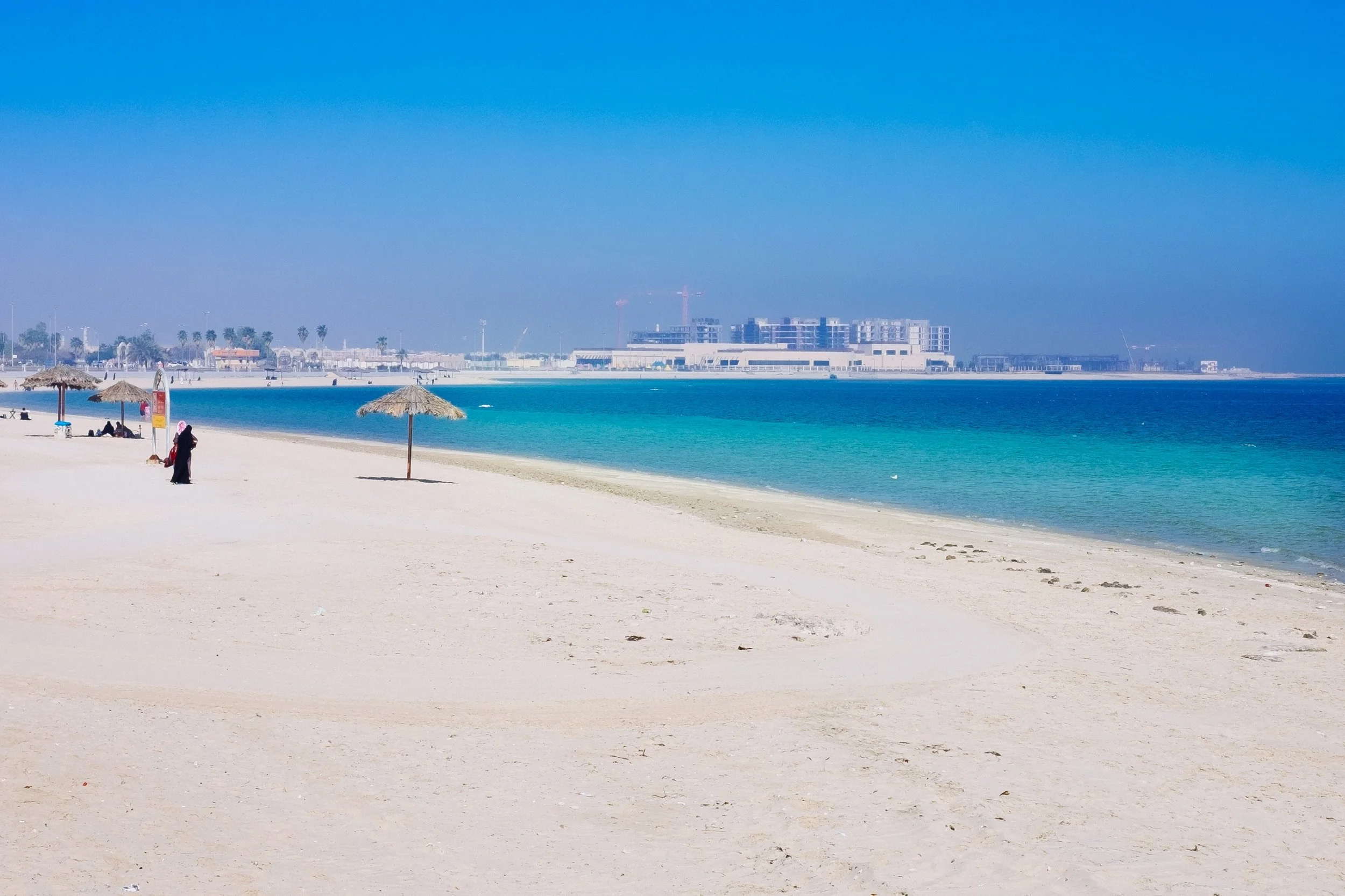 Scenic view of a beach with white sand, straw umbrellas, and a few people. The ocean has gradient shades of blue and turquoise. In the background, there are modern buildings and construction cranes under a clear blue sky.