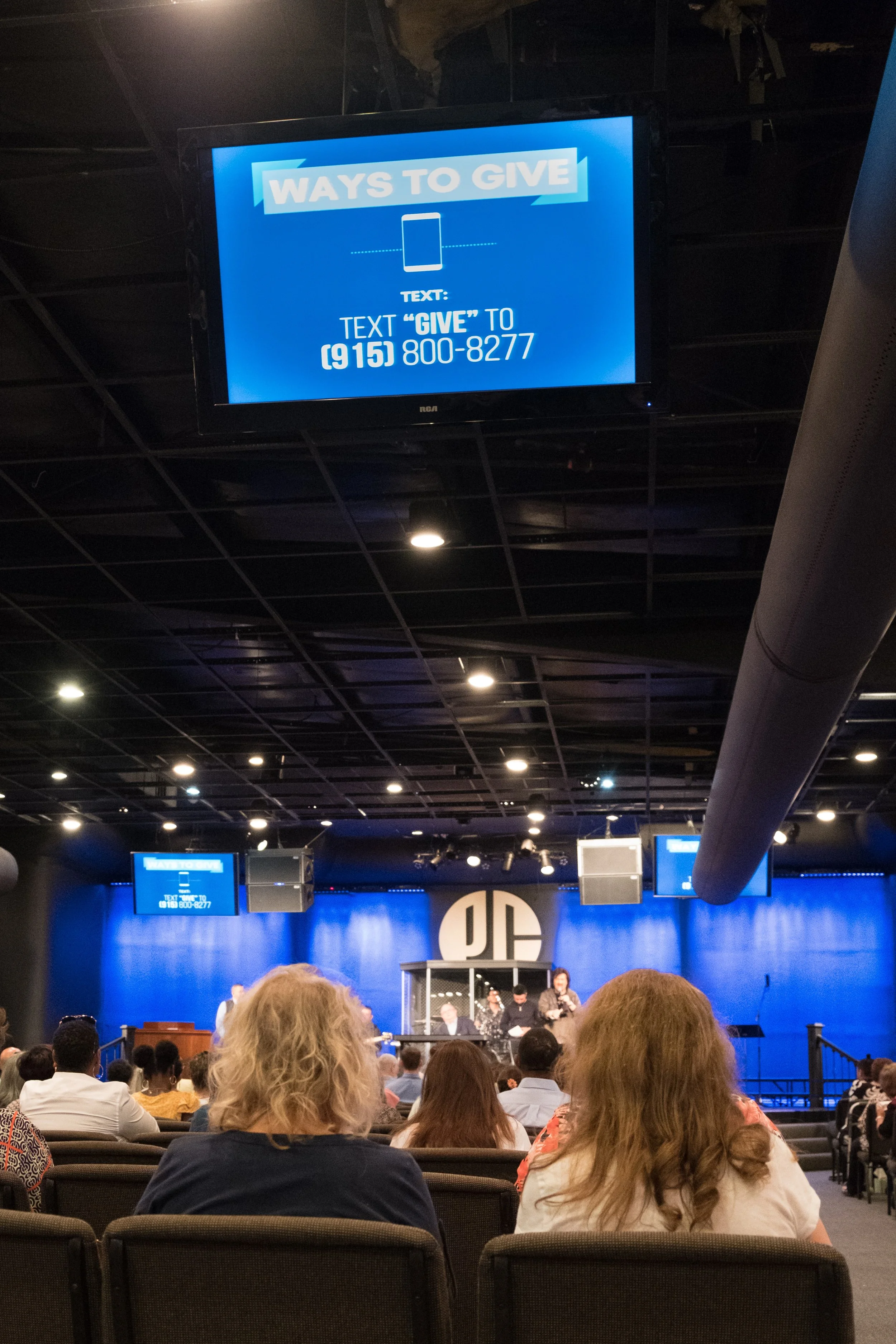 A screen displaying instructions on how to give money by texting 'GIVE' to a phone number in a large auditorium filled with people attending an event at DC Comics.