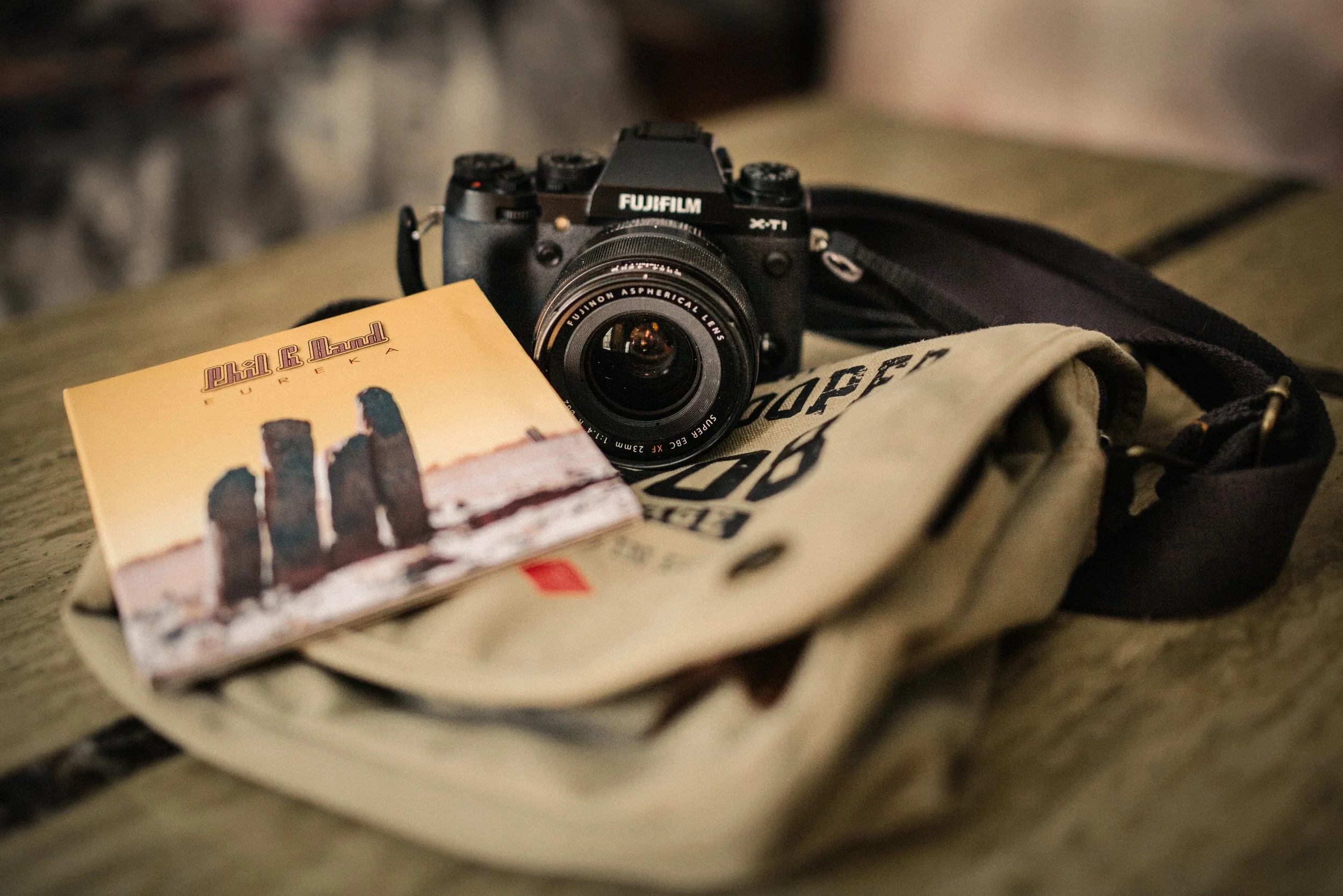 A Fujifilm X-series camera with a zoom lens resting on a beige bag, a book with a desert cover, and a patterned fabric background.