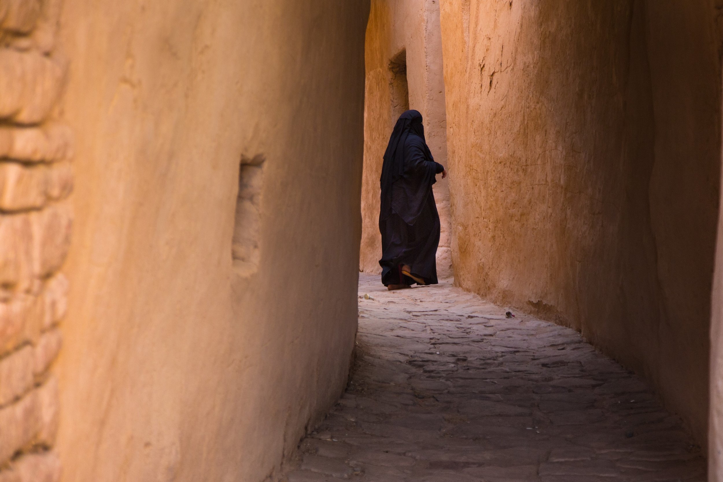 A person dressed in black clothing and head covering walking through a narrow alley with high beige stone walls.