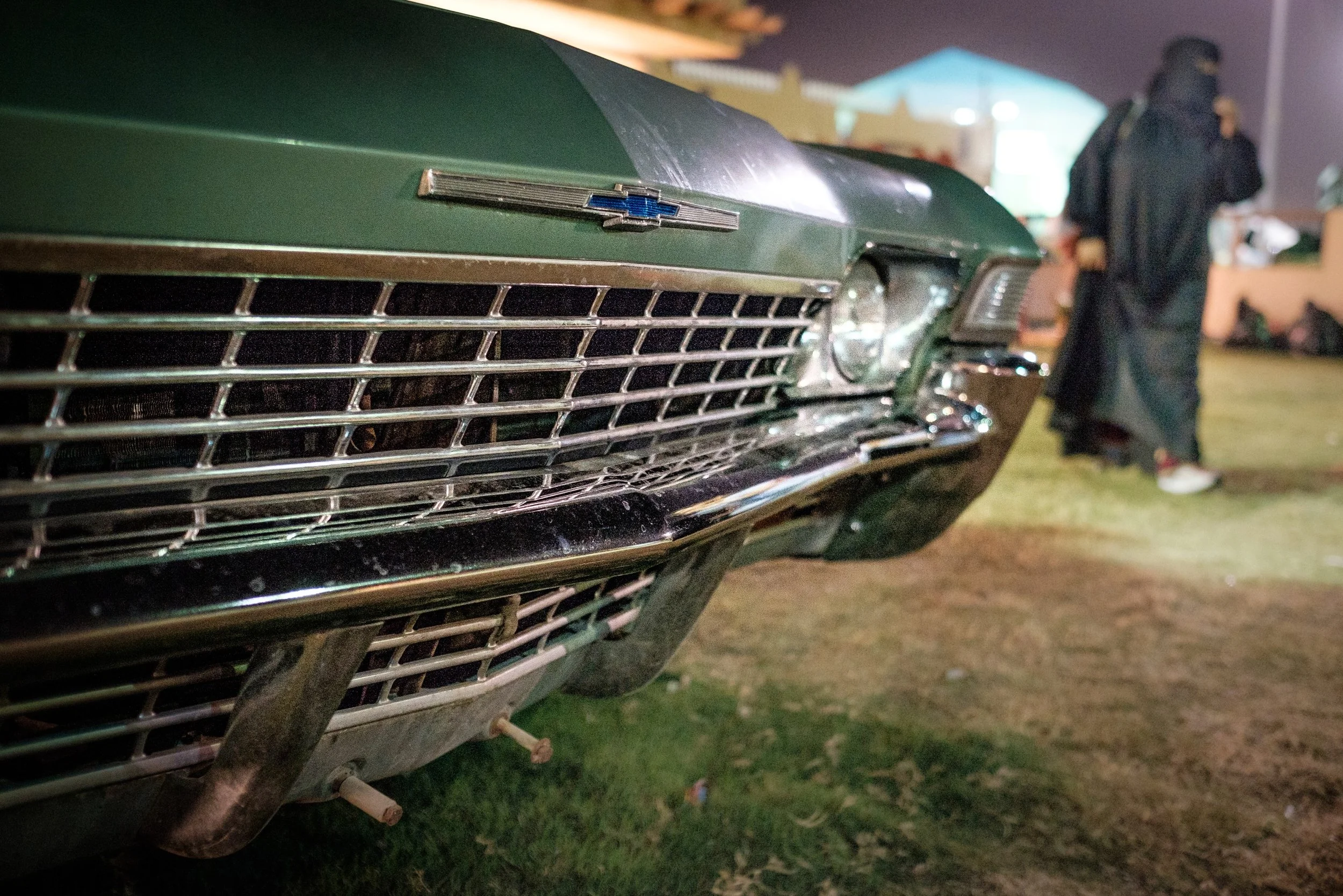 Close-up of the front grille of a green vintage Chevrolet car with a Chevrolet emblem, at night, with a person in dark clothing and a hat standing in the background.