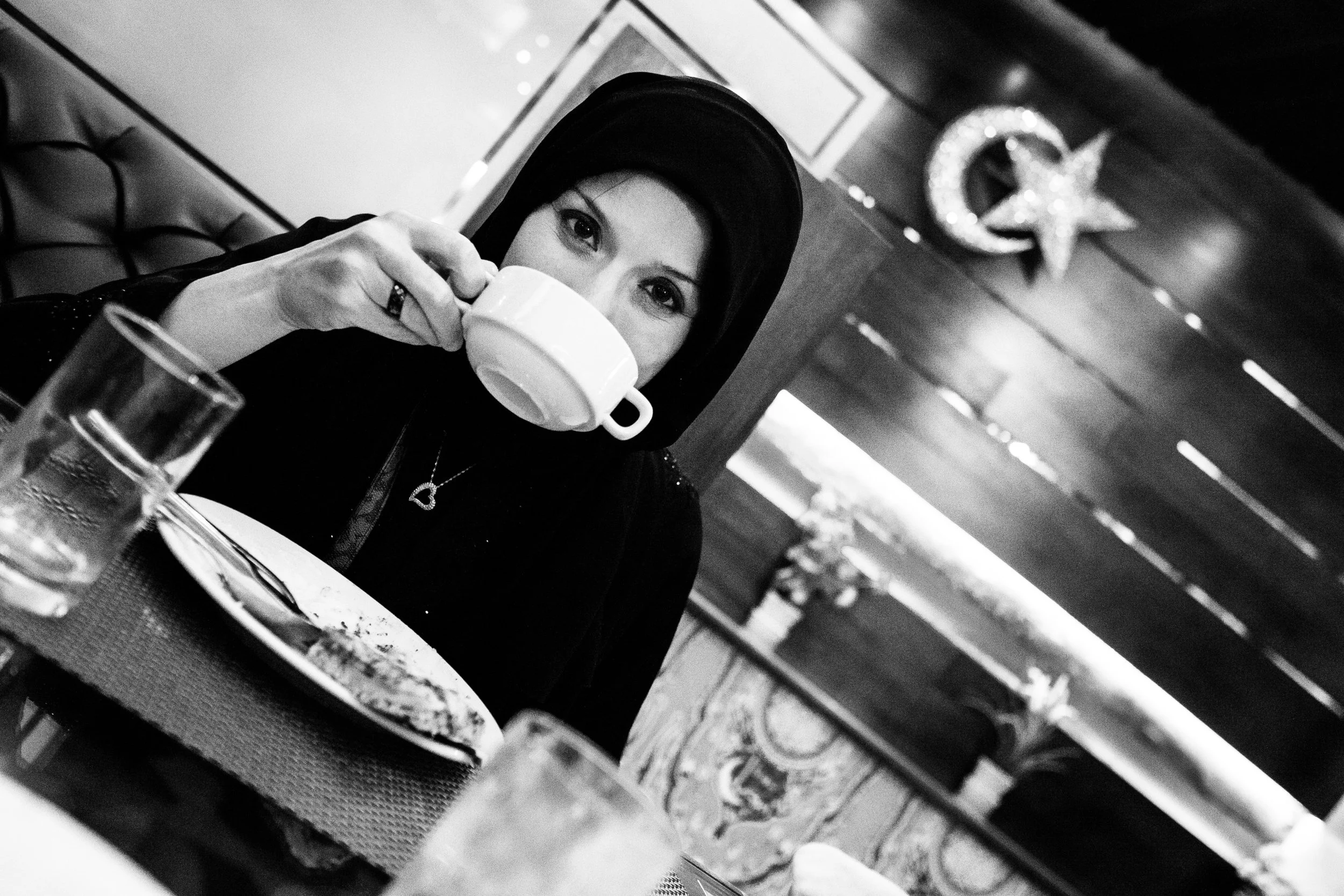 A woman with dark hair wearing a hat and dark clothing is sitting at a table, drinking from a white mug in a restaurant or cafe setting. There is a glass of water and a plate with some food in front of her. The background has decorations, including a