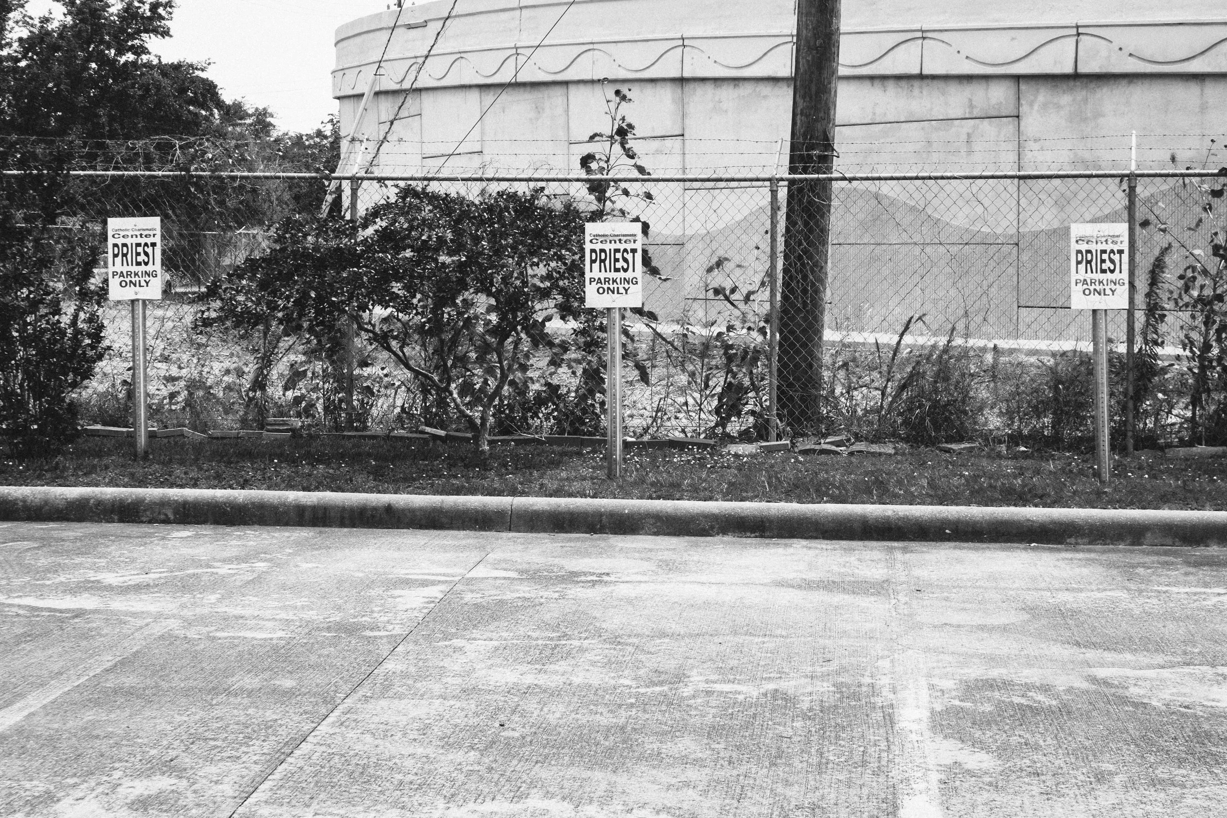 Black and white photo of a parking lot with a chain-link fence and signage reading 'Priest Parking Only.' Plants grow along the fence, and there is a large building or structure in the background.