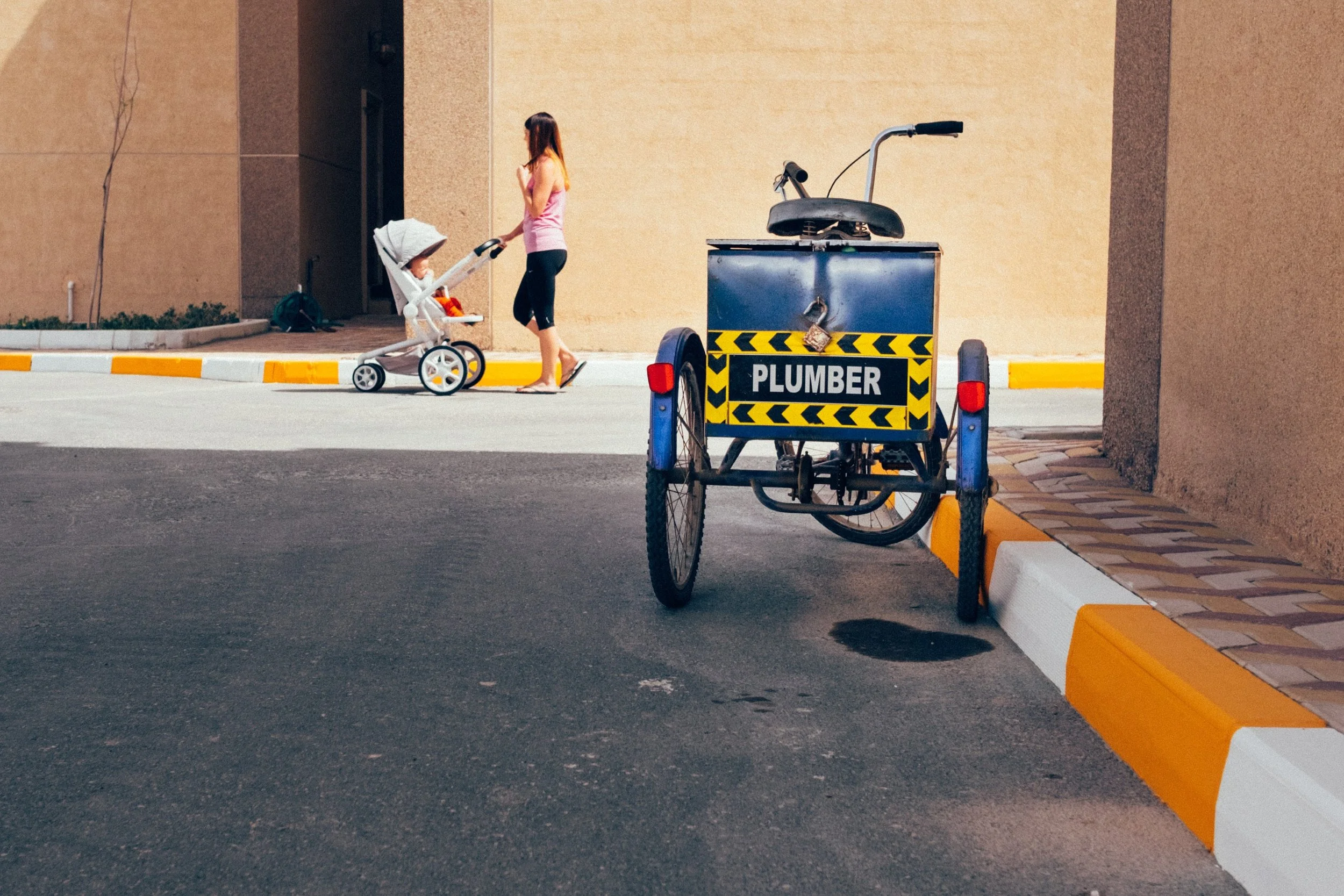 A woman walking on the sidewalk pushing a stroller with a baby, next to a blue plumbing cart with yellow and black caution tape.