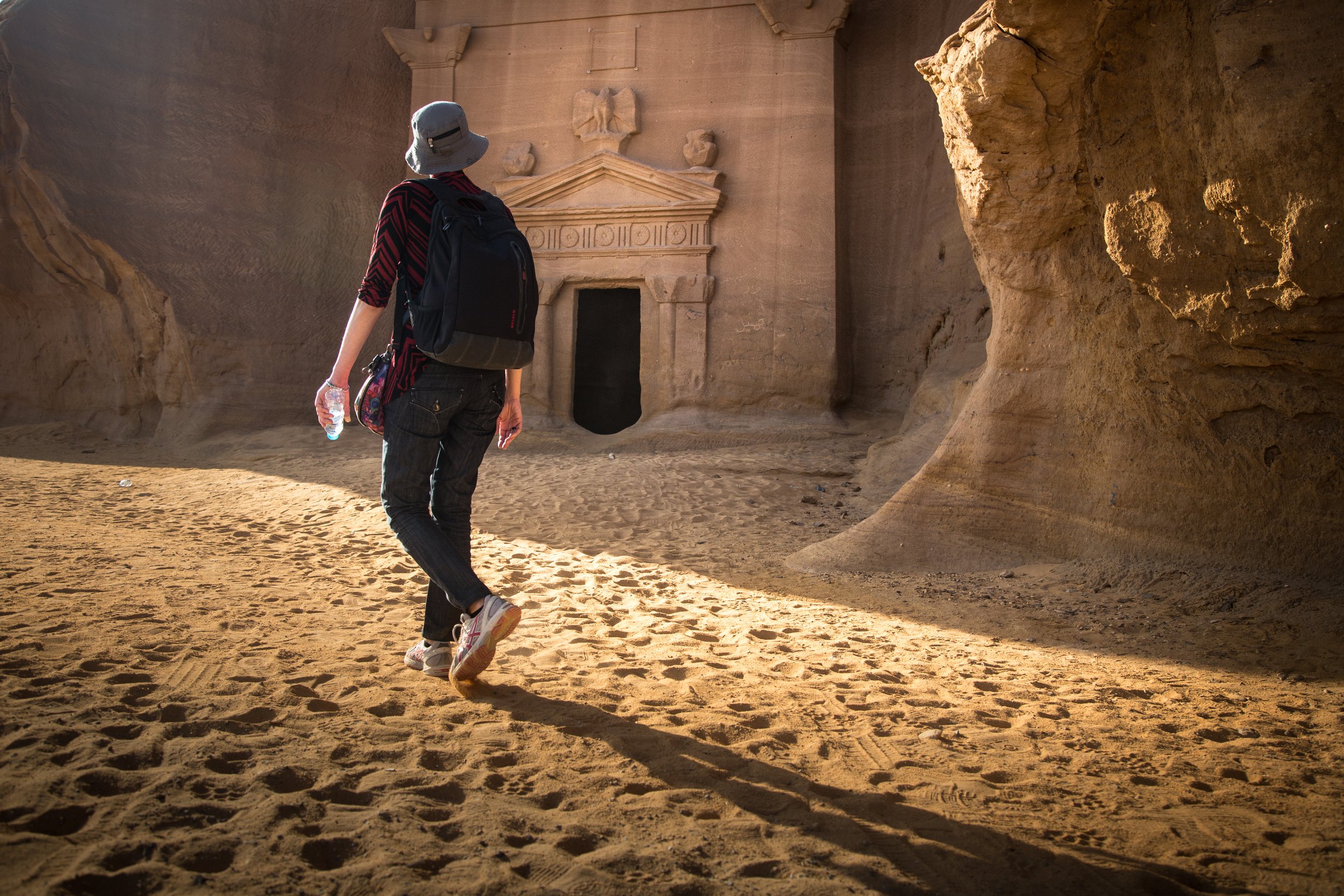 Person walking through a rocky desert area with ancient carved stone structure in the background, holding a water bottle.