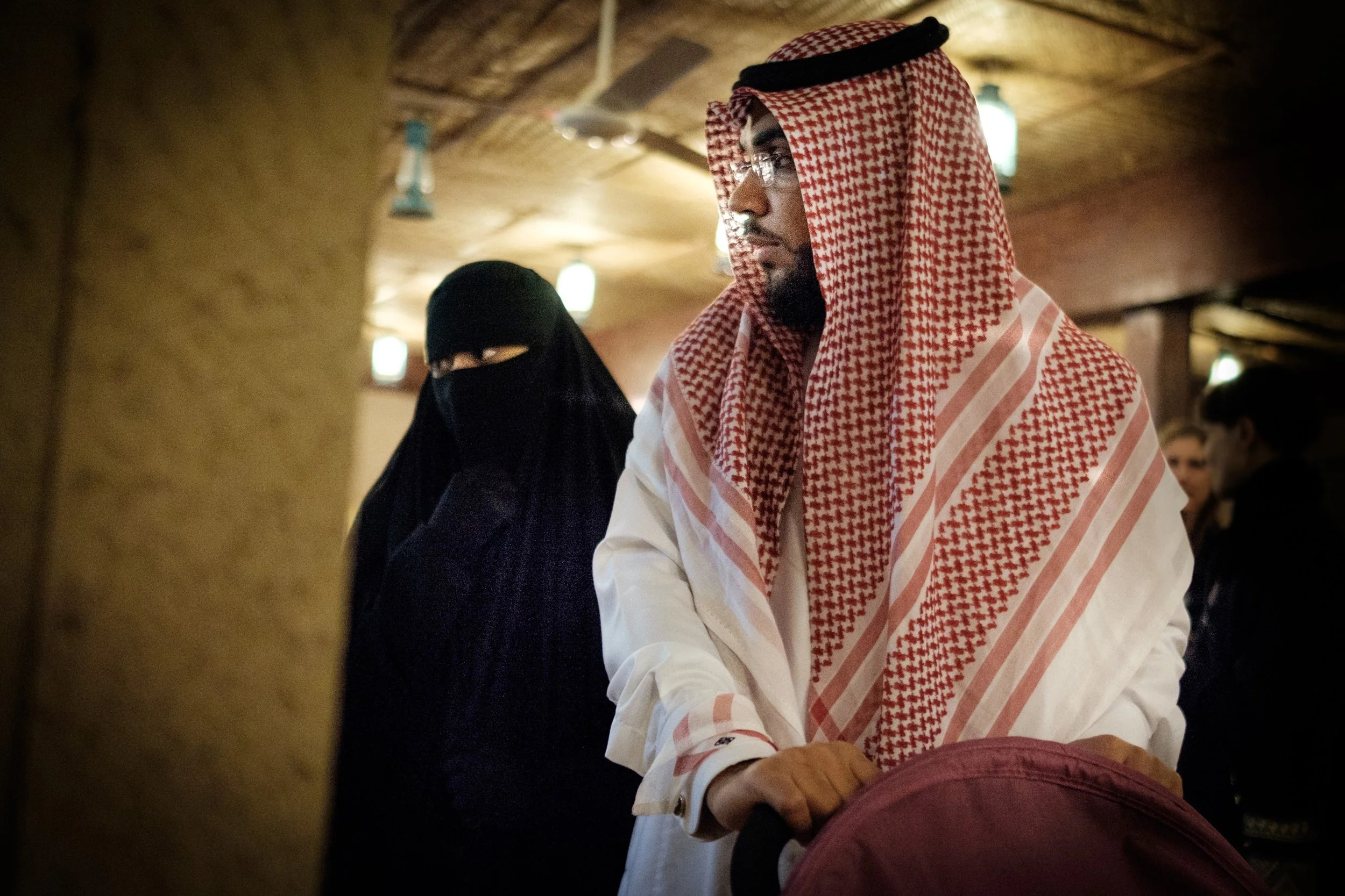 A man dressed in traditional Middle Eastern attire, wearing a red and white keffiyeh and white robe, stands in a dimly lit room with wooden beams on the ceiling. He is looking down and slightly to the side, with several people and warm lighting in th