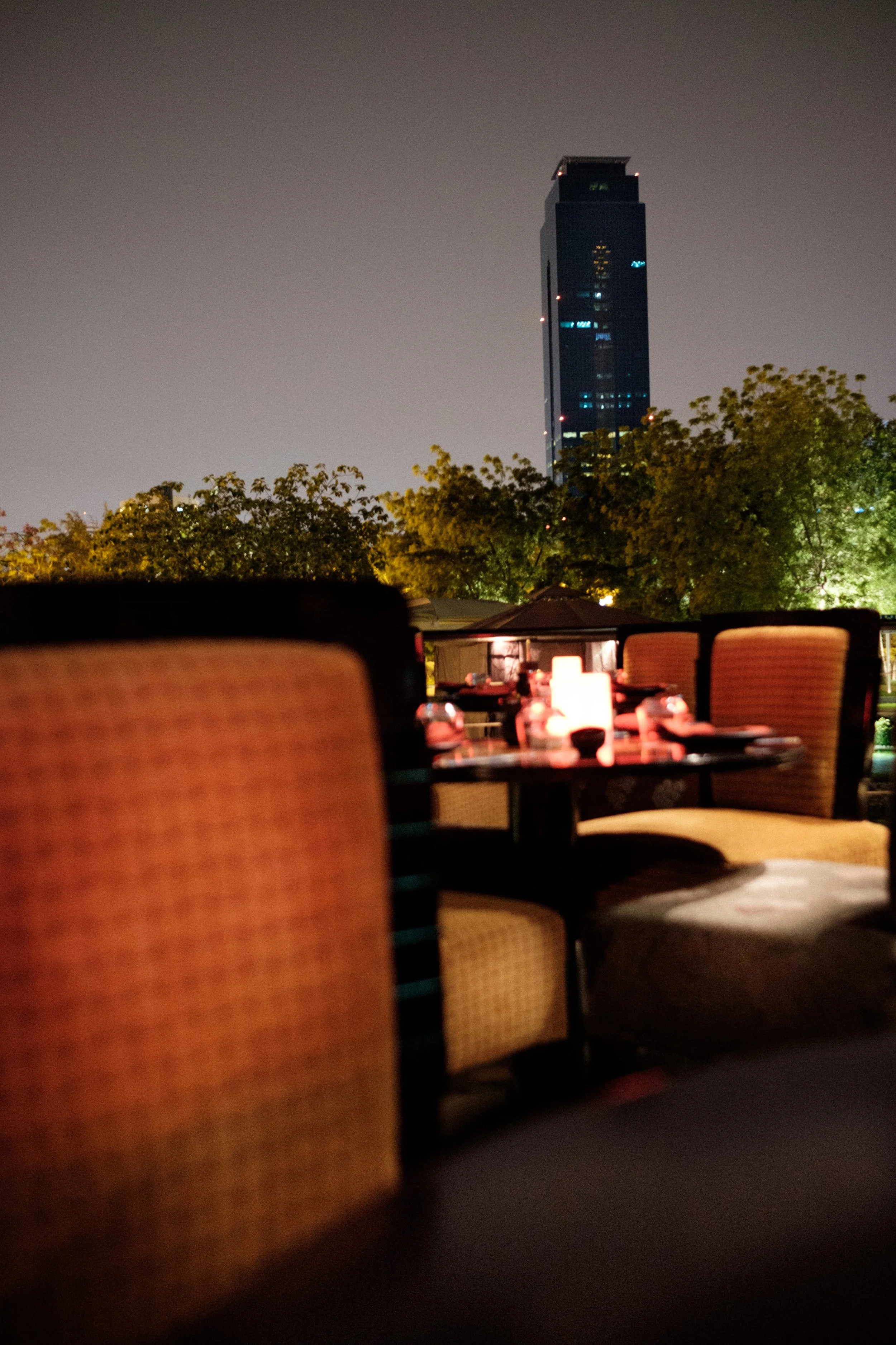 Night view of a cityscape with a very tall dark building illuminated with lights, trees in the foreground, and an outdoor dining area with tables and chairs.