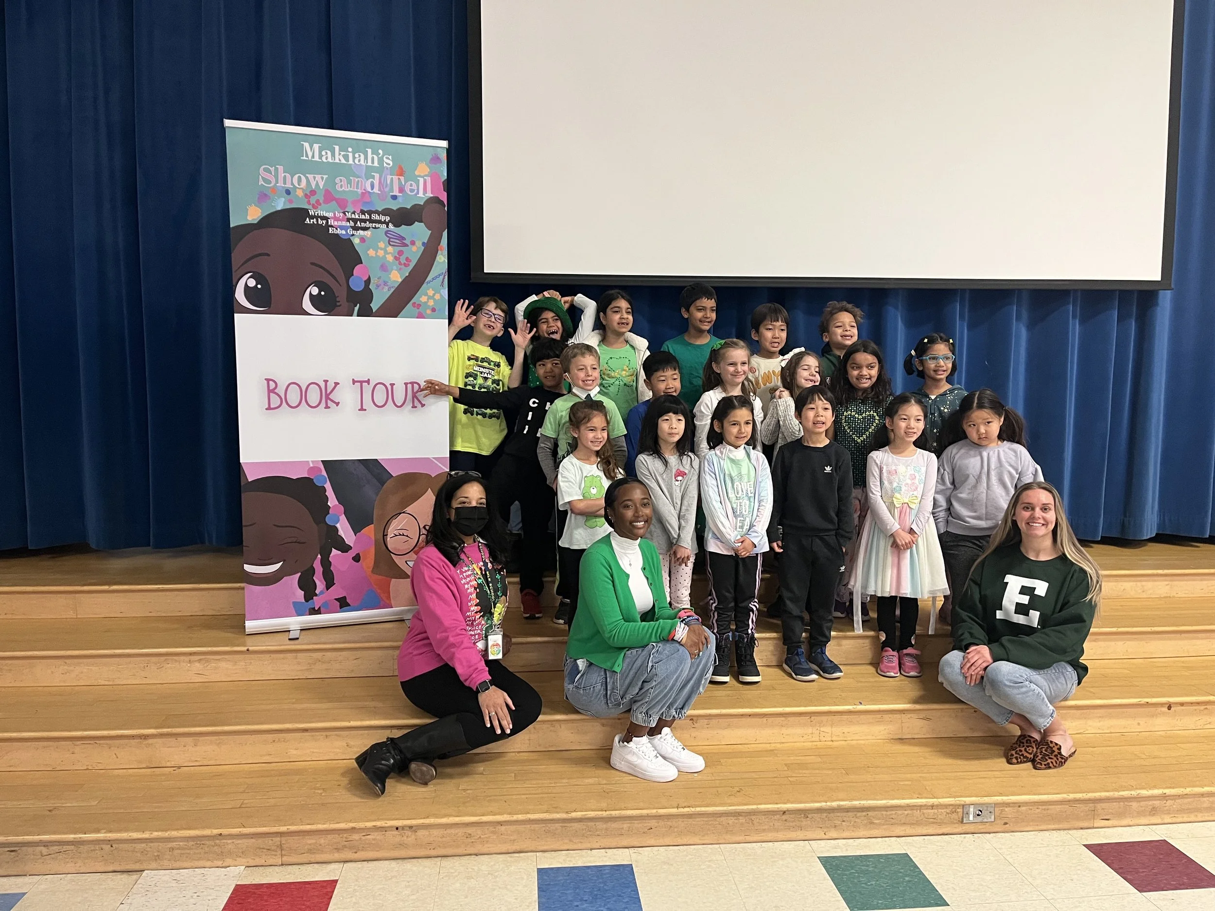 Group of children and adults on stage for a school event, with a colorful banner that reads 'Makah's Show and Tell Book Tour'.