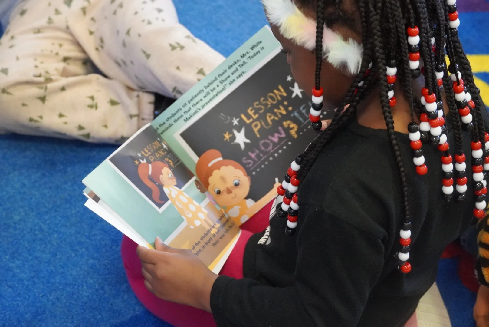 A young girl with braided hair and a furry animal ear headband sitting on a pink cushion, reading a colorful children's book, called Makiah's Show and Tell by Makiah Corum, with illustrations of two animated characters on the pages.