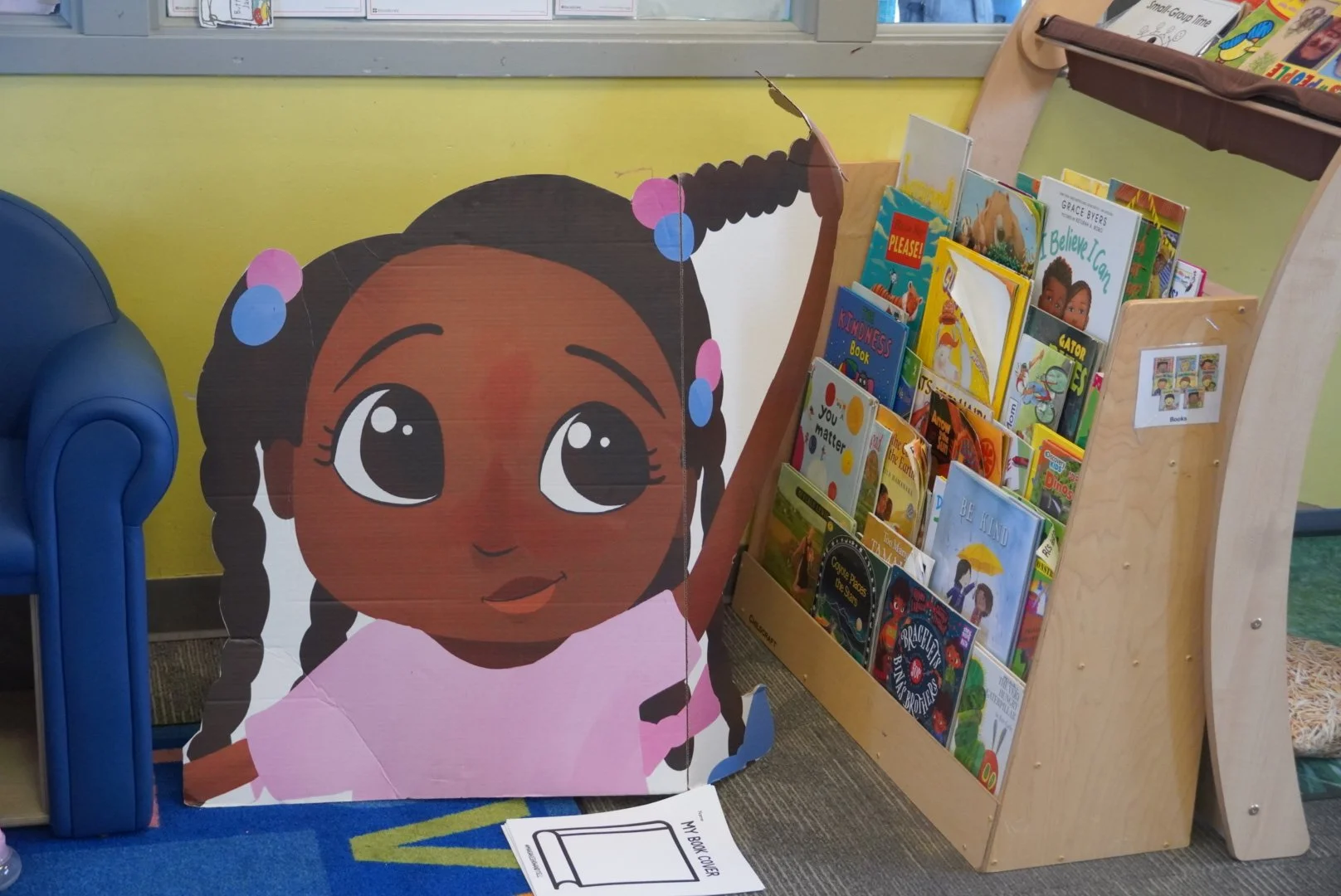 A large cartoon cutout of a girl with dark hair in braids, decorated with pink and blue hair ties, and wearing a pink shirt. Next to it is a small wooden bookshelf filled with children's books.