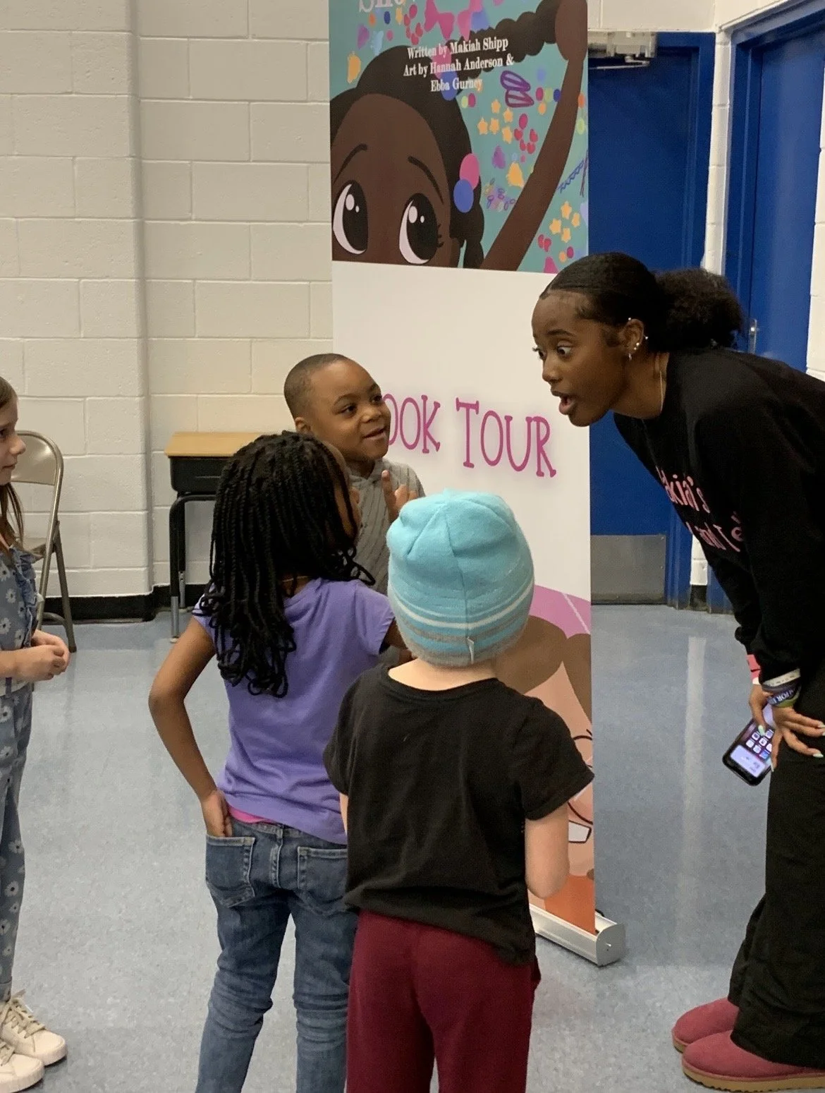 A group of children and an adult woman talking in front of a colorful book tour sign with artwork of a girl with big eyes and hairstyle, inside a room with light-colored brick walls and a blue door.