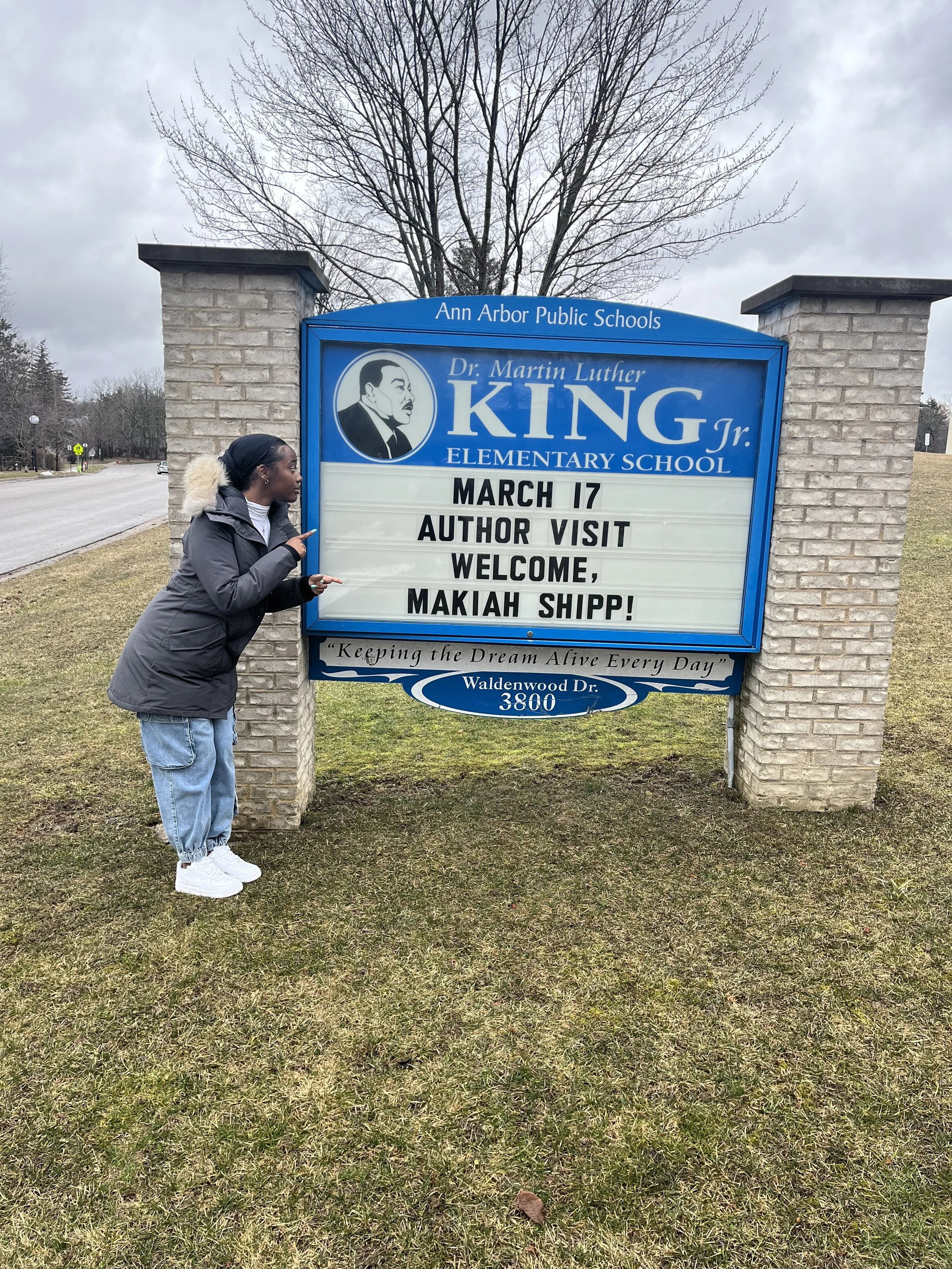 A woman standing next to a school sign with a portrait of Dr. Martin Luther King Jr. The sign announces a visit from the author Makiah Shipp on March 17 at Ann Arbor Public Schools.