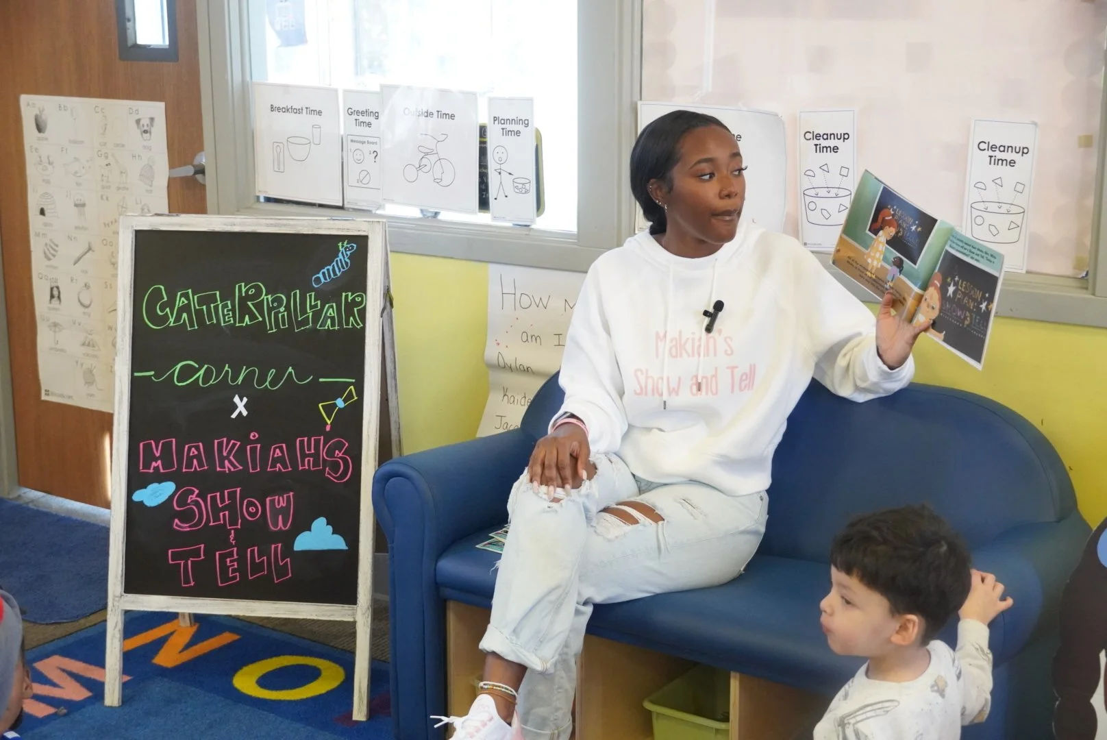 Young woman reading a children's book to a group in a classroom setting. She sits on a blue sofa, wearing a white hoodie and ripped jeans. A blackboard sign next to her displays 'Caterpillar Corner x Maki's Show & Tell'. Several children are seated nearby, attentive. Classroom hyper-visual decor includes educational posters and signs with time schedules.