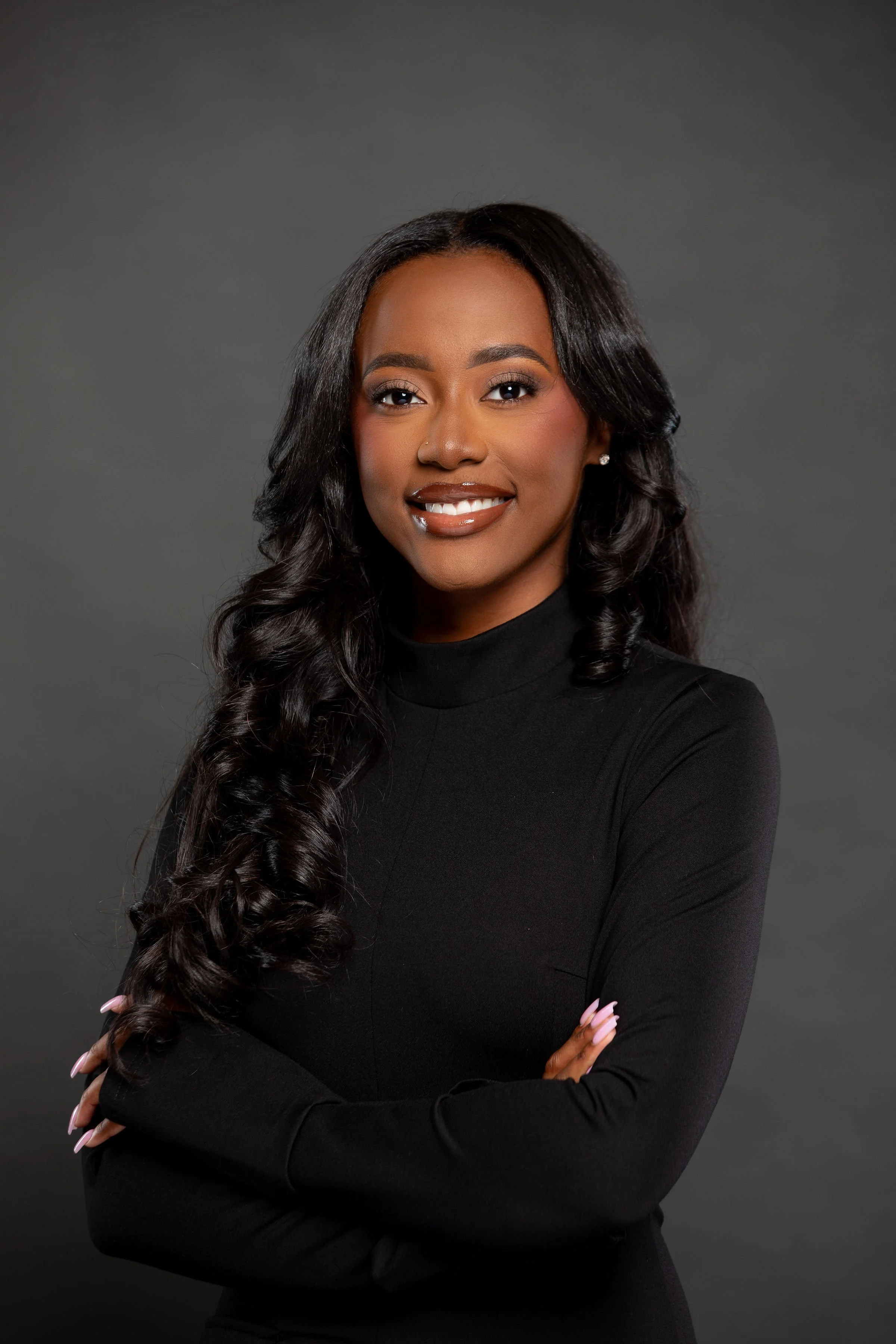 Portrait of a Black woman with long, curly black hair, wearing a black turtleneck, smiling, with arms crossed against a gray background.