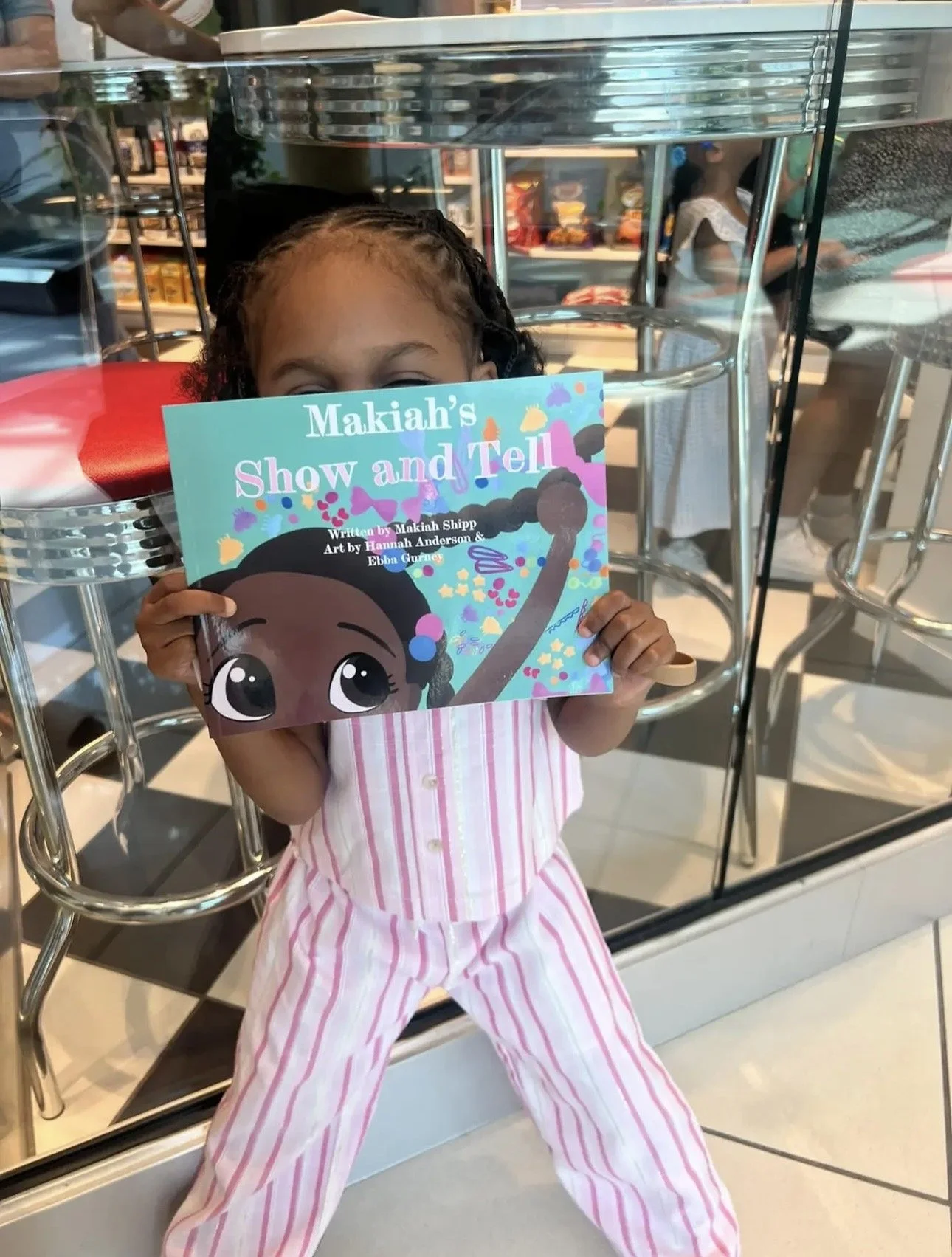 A young girl  is standing in a store, holding a colorful book titled "Makiahs Show and Tell," with only her eyes and forehead visible peeking over the top of the book.