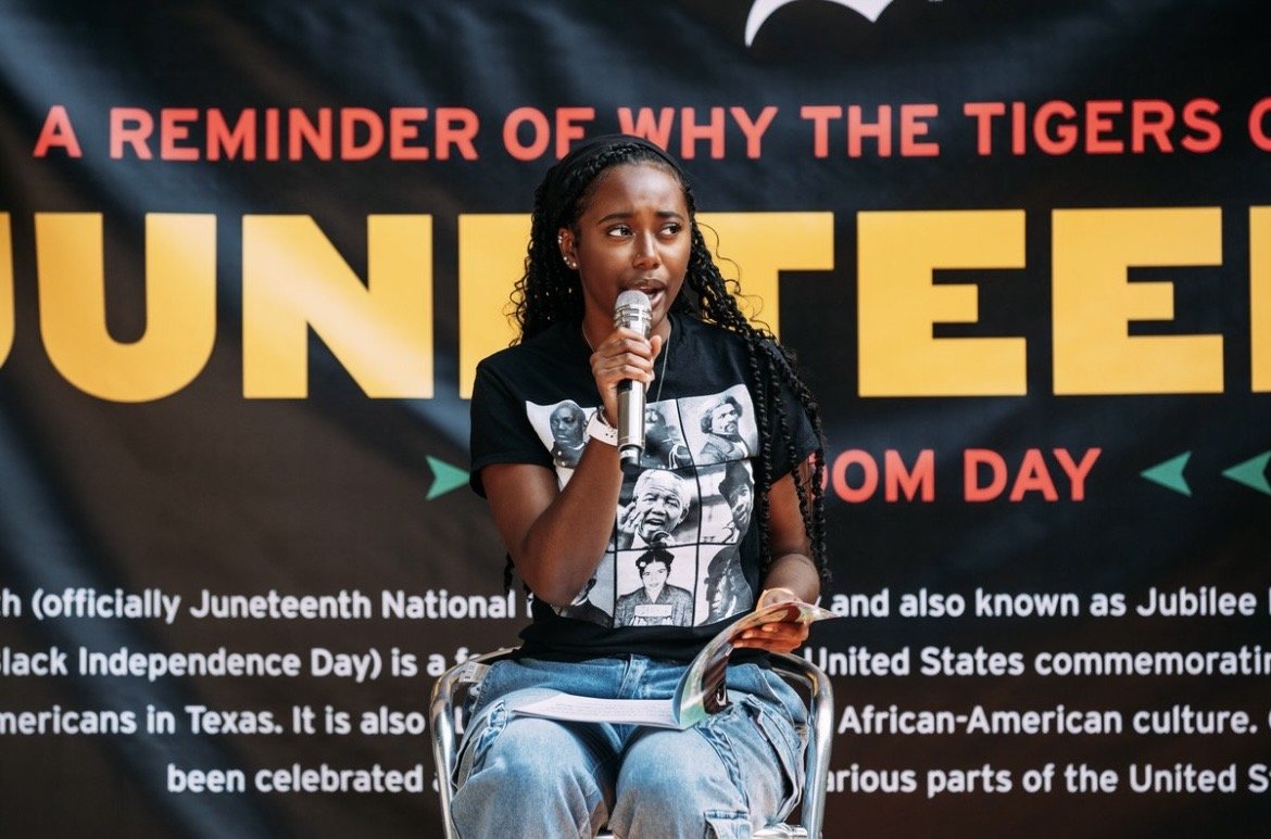 Young girl speaking into a microphone at Juneteenth celebration event, with a black background banner displaying the words 'A REMINDER OF WHY THE TIGERS' and 'JUNETEENTH' in large yellow letters, and smaller text about Juneteenth and African-American culture.