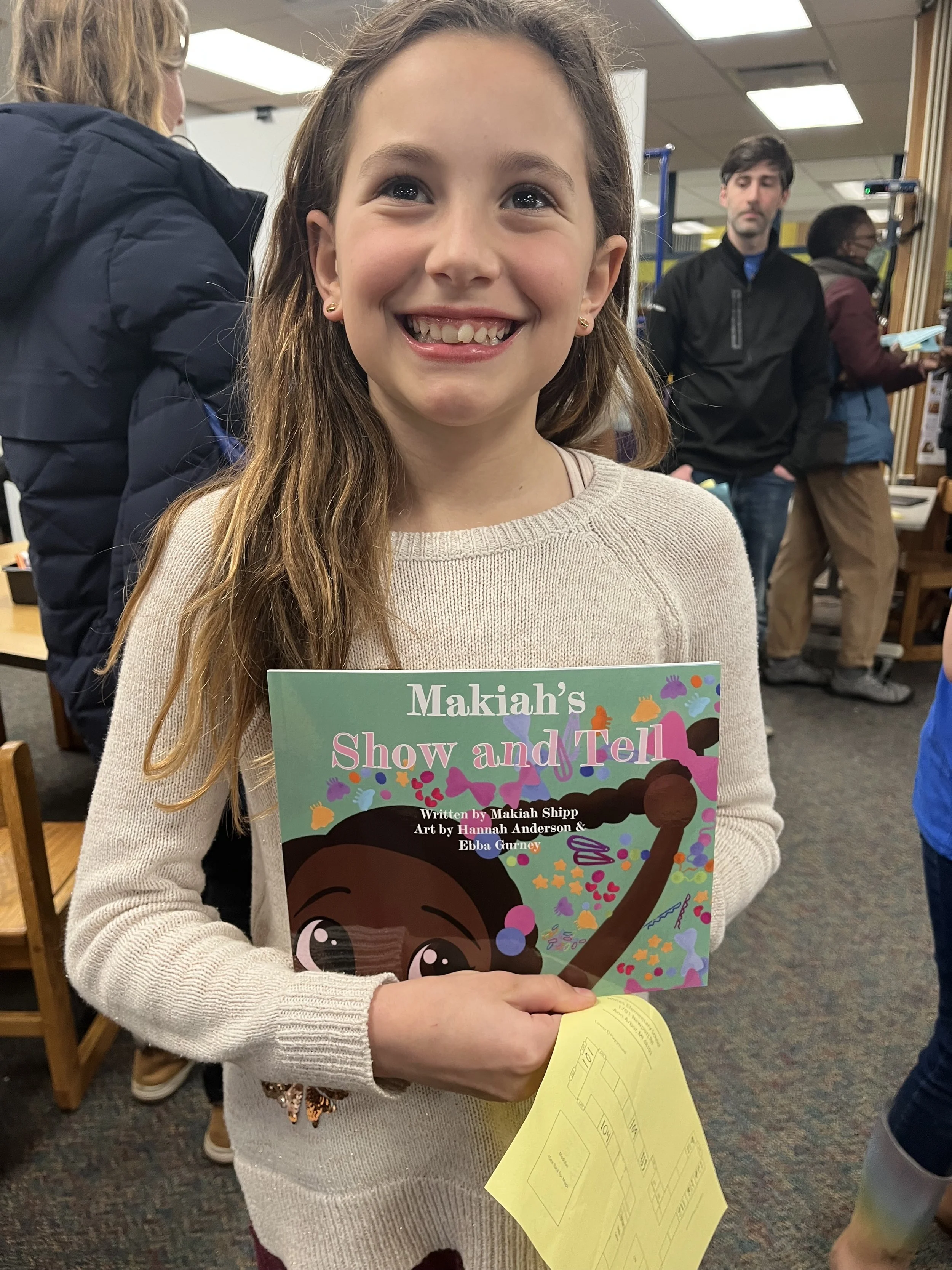 A young girl smiling and holding a children's book titled 'Makiahs Show and Tell' with colorful illustrations on the cover. She is in a room with other people and appears to be at a school or bookstore event.