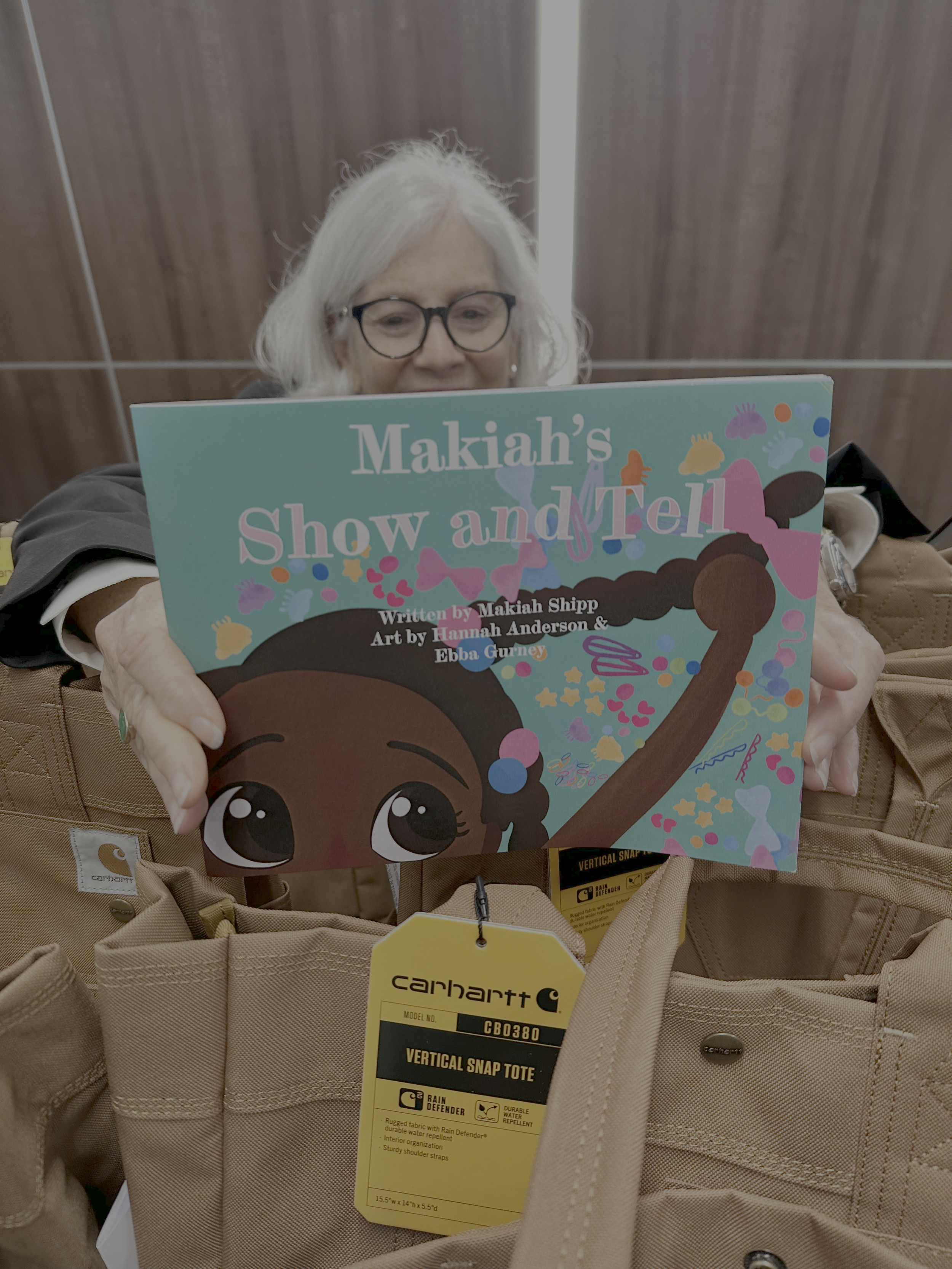 A woman with glasses and white hair is holding a colorful children's book titled "Makiahs Show and Tell" in front of her face. She is surrounded by brown tote bags and is in an indoor setting.