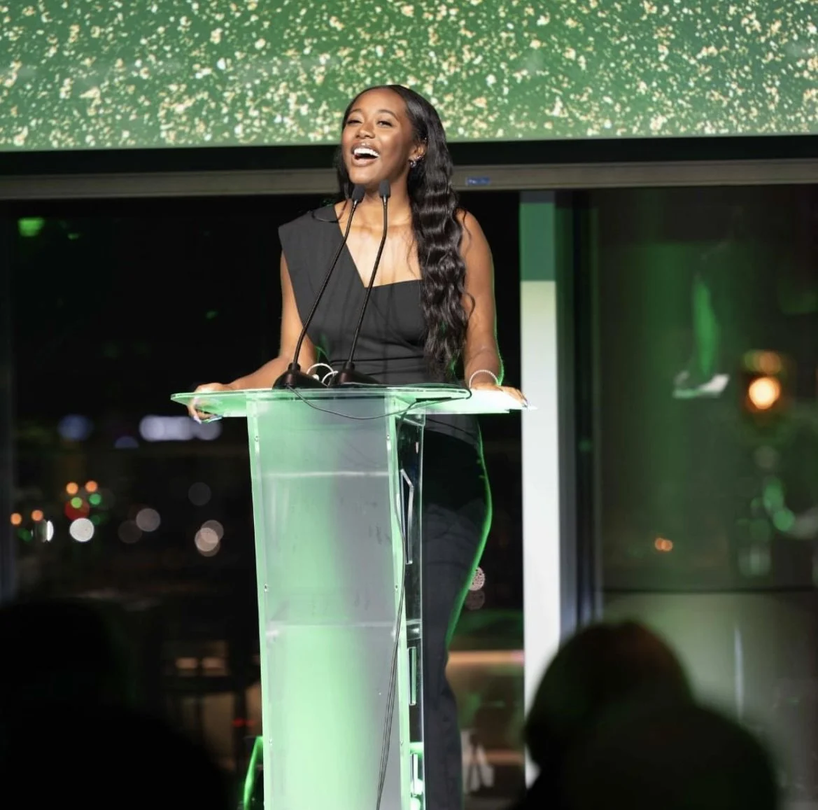 A woman with long black wavy hair, wearing a sleeveless black dress, standing at a clear podium with microphones, smiling and speaking at an event with a large screen behind her.