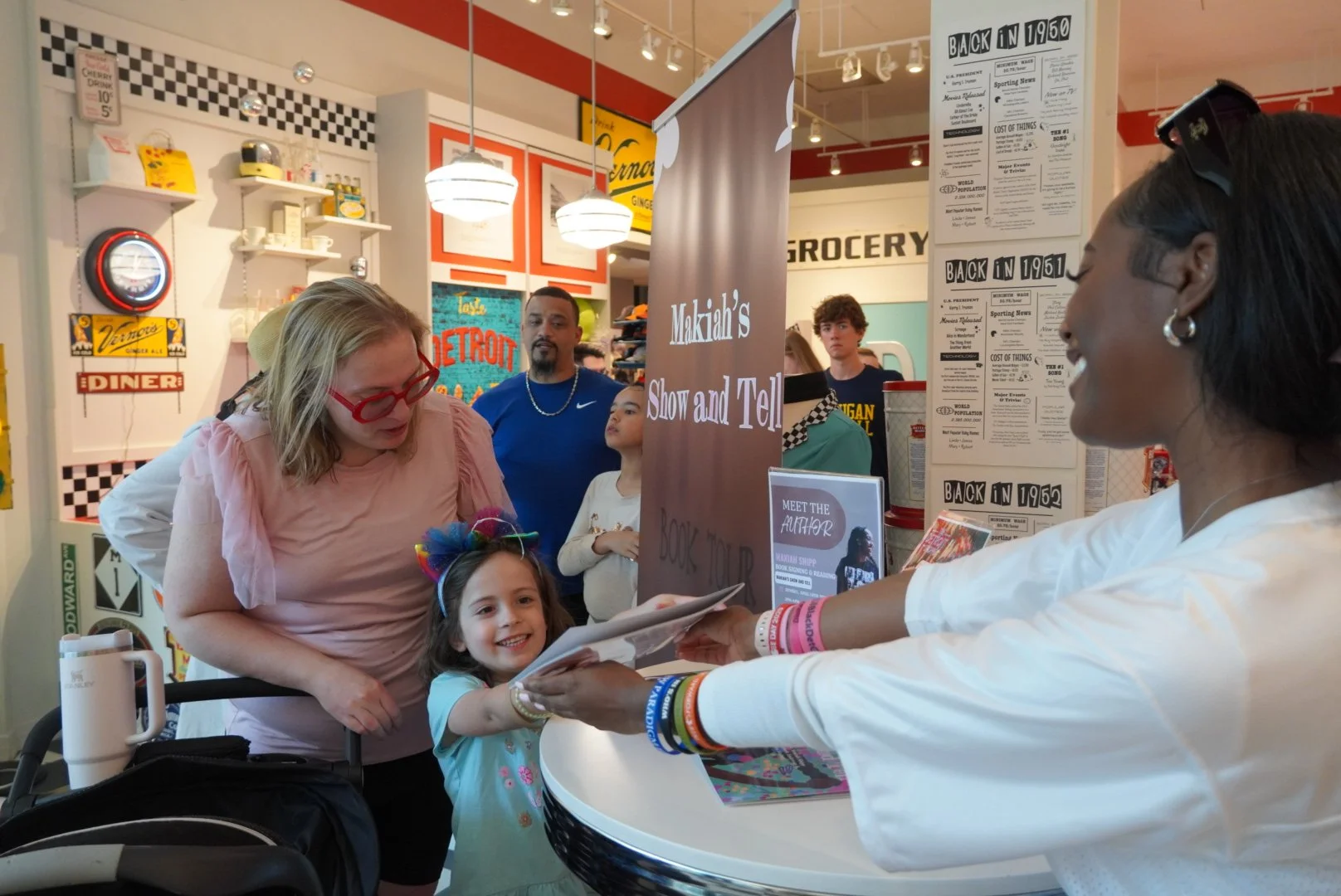 A young girl smiling and receiving a book from a woman at a book signing event. Several people are gathered around, and there are vintage-themed decorations in the background.