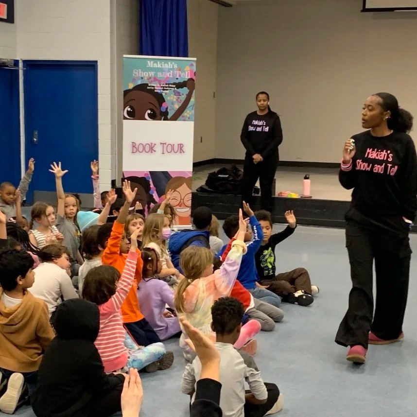 A group of children sitting on the floor in a room, raising their hands during a book tour presentation by two women in black sweatshirts with pink and purple text, in front of a colorful sign that reads 'Makah's Show and Tell' and 'Book Tour'.