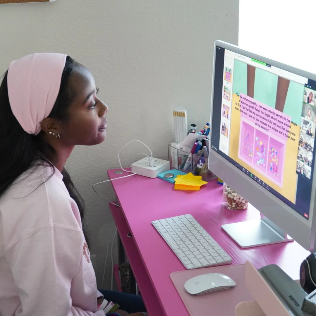 Girl with long dark hair and a pink headband sitting at a pink desk, looking at a large computer monitor displaying a colorful animation and text.