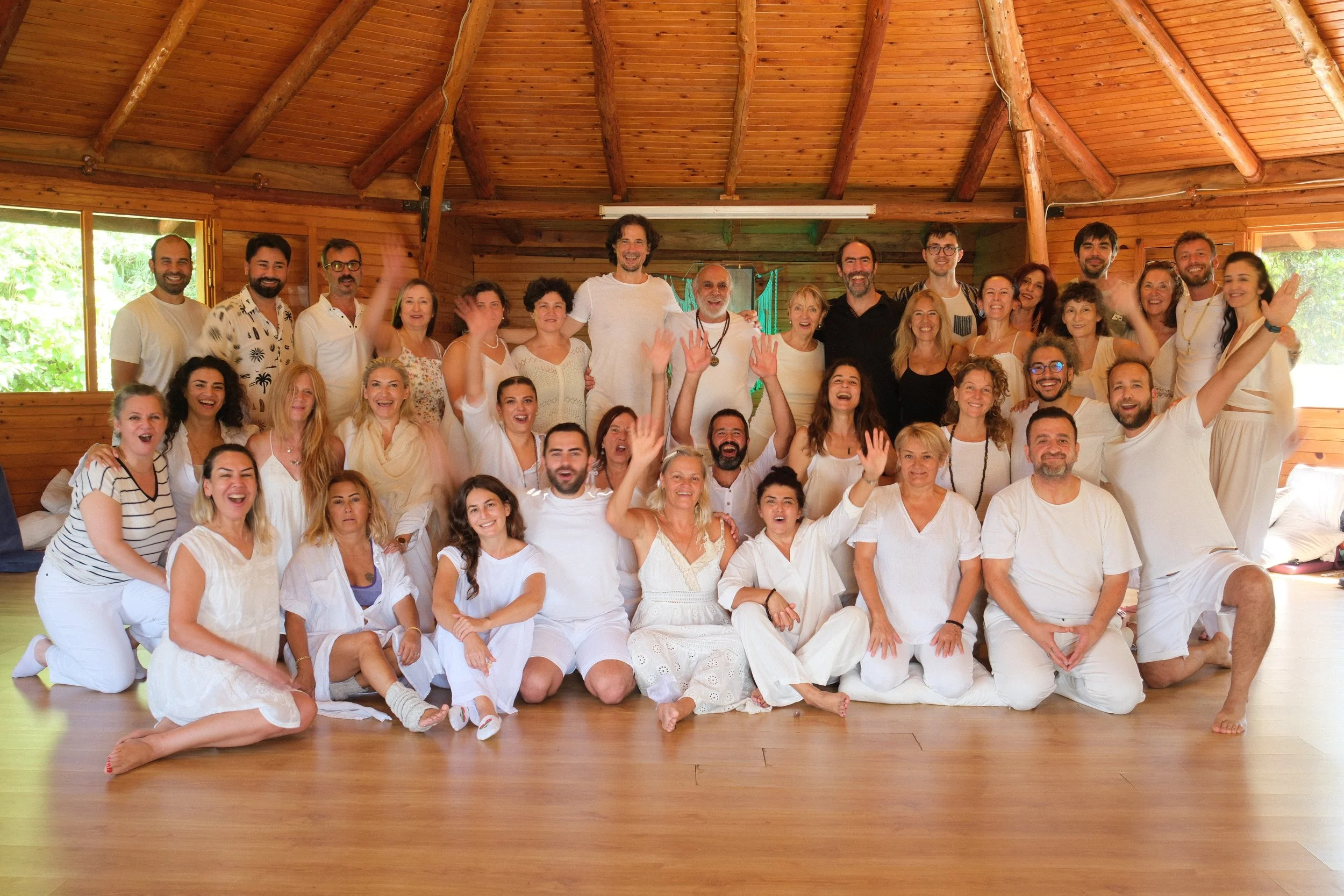 Group of smiling people dressed in white, inside a wooden cabin, some sitting on the floor and others standing, celebrating together. Osho celebration, evening meeting, diamond breath school.