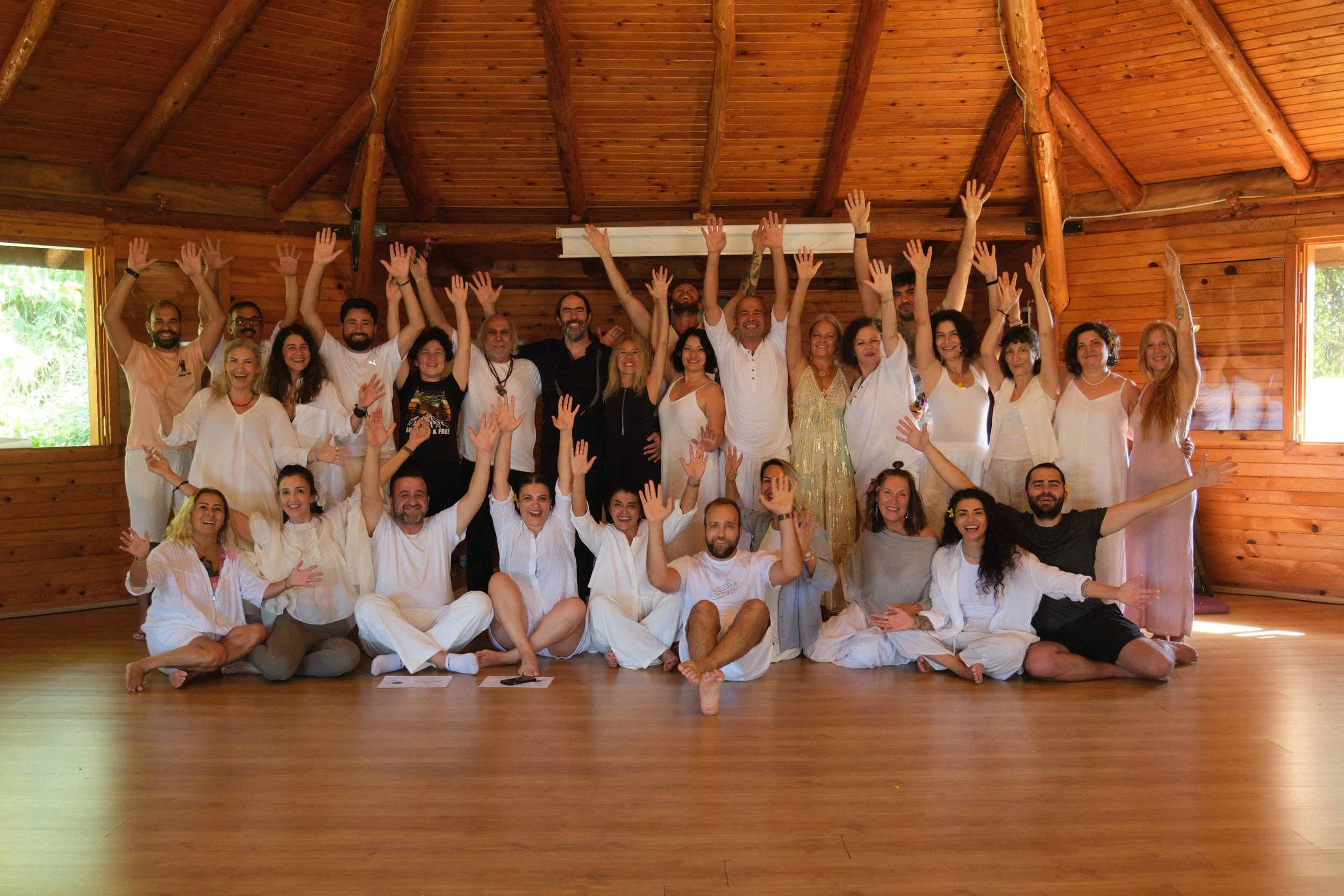 A large group of people, smiling and raising their hands, gathered in a wooden room with natural light coming through the windows. Osho celebration, evening meeting, diamond breath school.