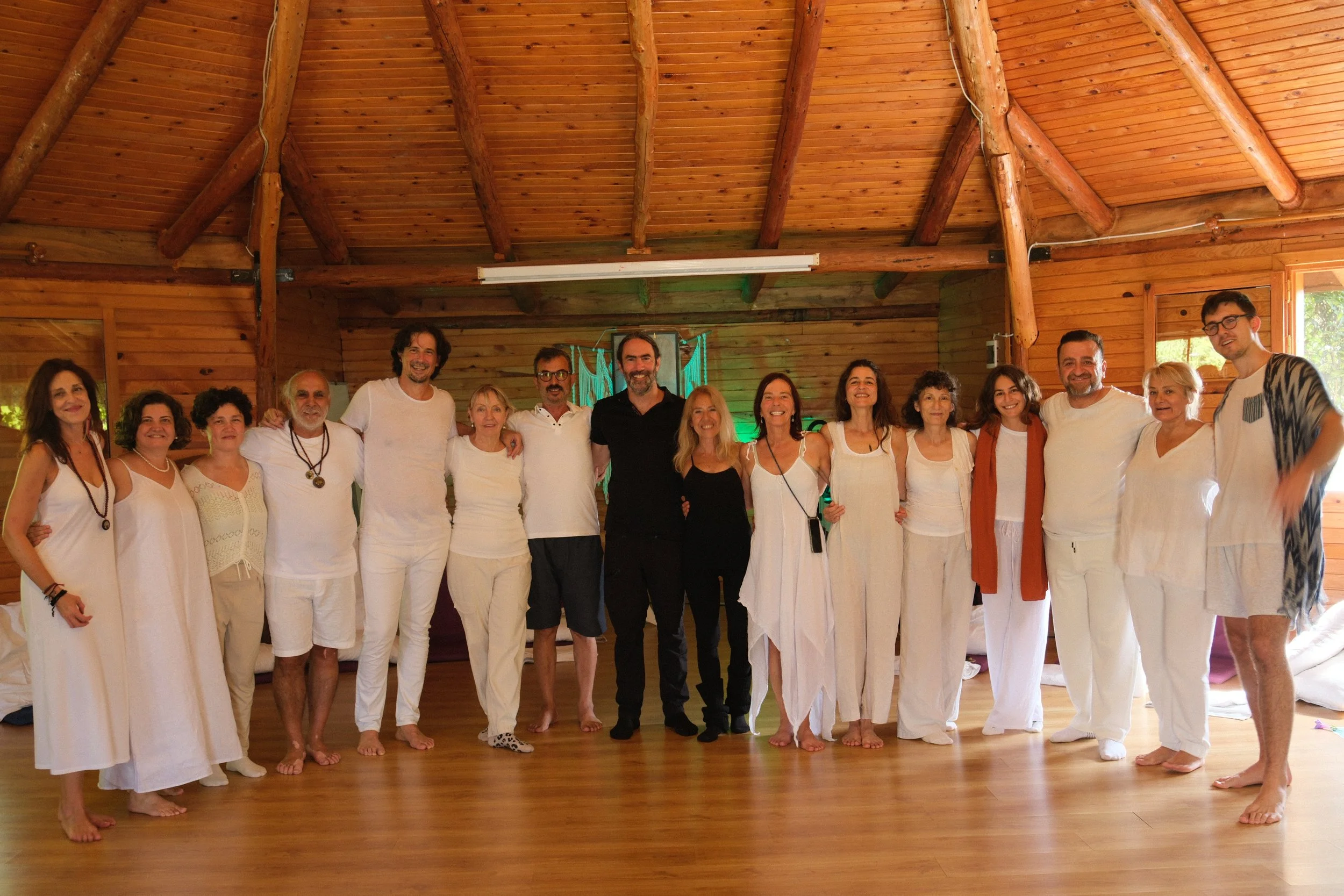 Group of people standing together inside a wooden structure, smiling. Some individuals are dressed in white, barefoot, and the setting appears casual and warm. Osho celebration, evening meeting, diamond breath school.