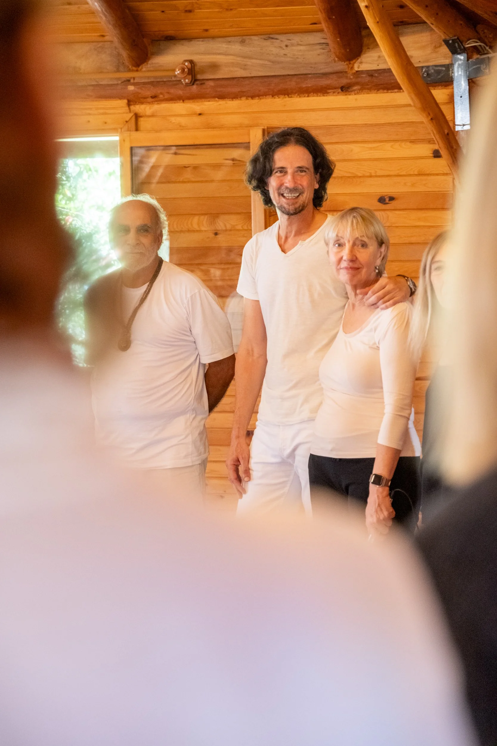 Smiling man with dark curly hair and beard, wearing a white V-neck T-shirt, standing next to a older woman with short blonde hair and earrings, in a wooden room with a window in the background.