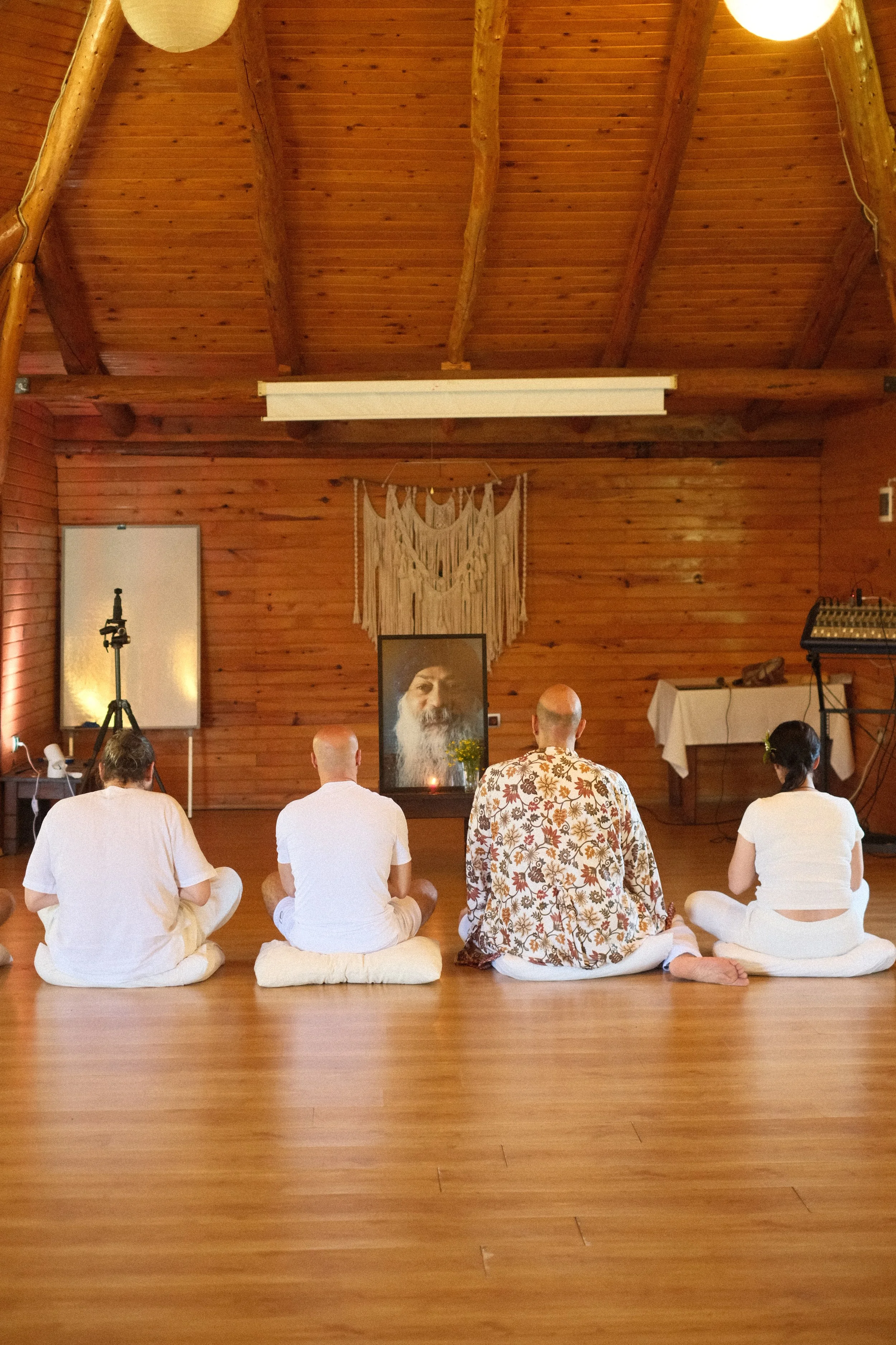 People in white clothing sitting cross-legged on cushions in a wooden room, facing a portrait of a bearded man and a small altar, participating in a meditation or spiritual ceremony. Osho celebration, evening meeting, diamond breath school.