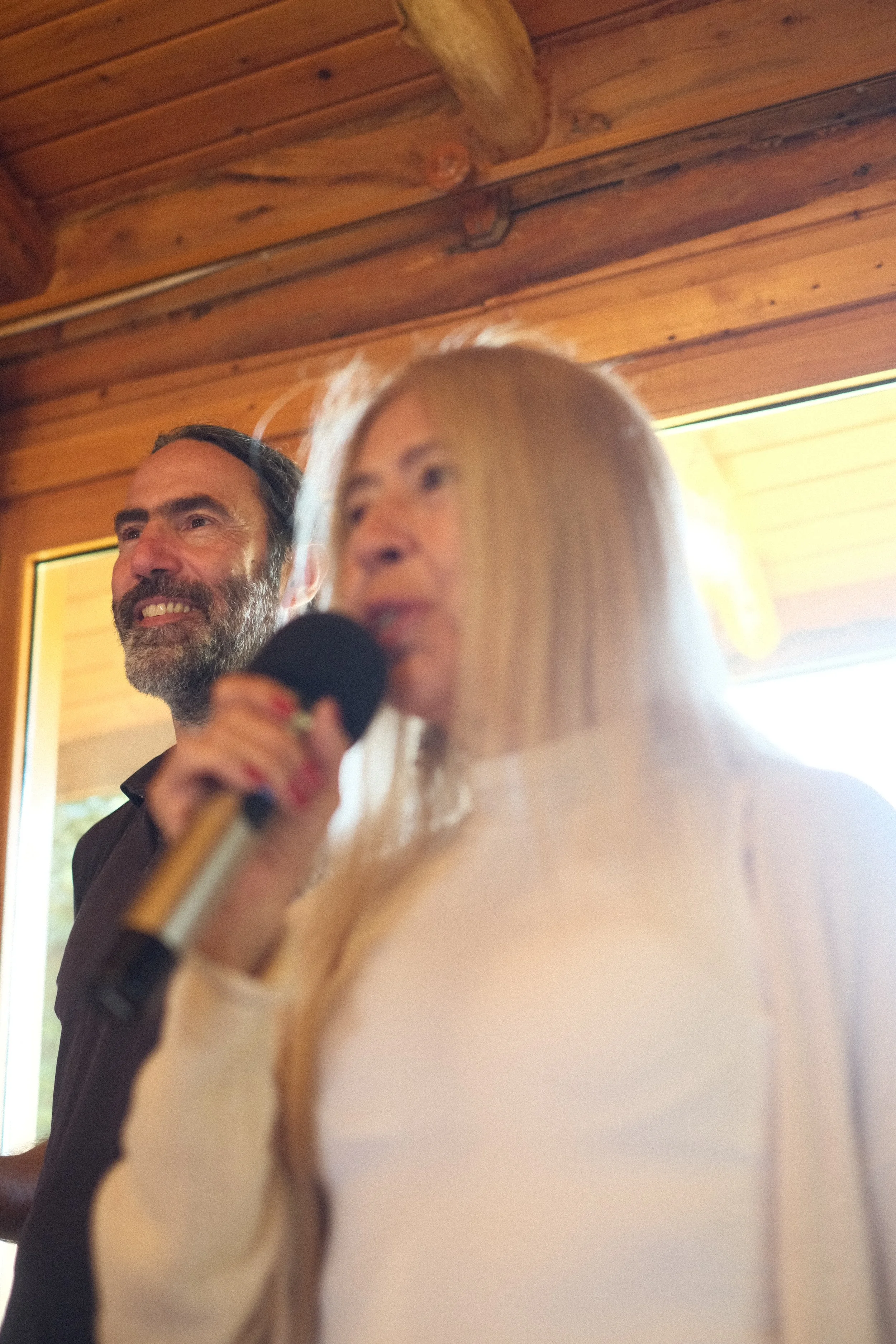 A man with a beard and a woman holding a microphone, both near a wooden ceiling with large beams, in a cozy indoor setting with natural light.