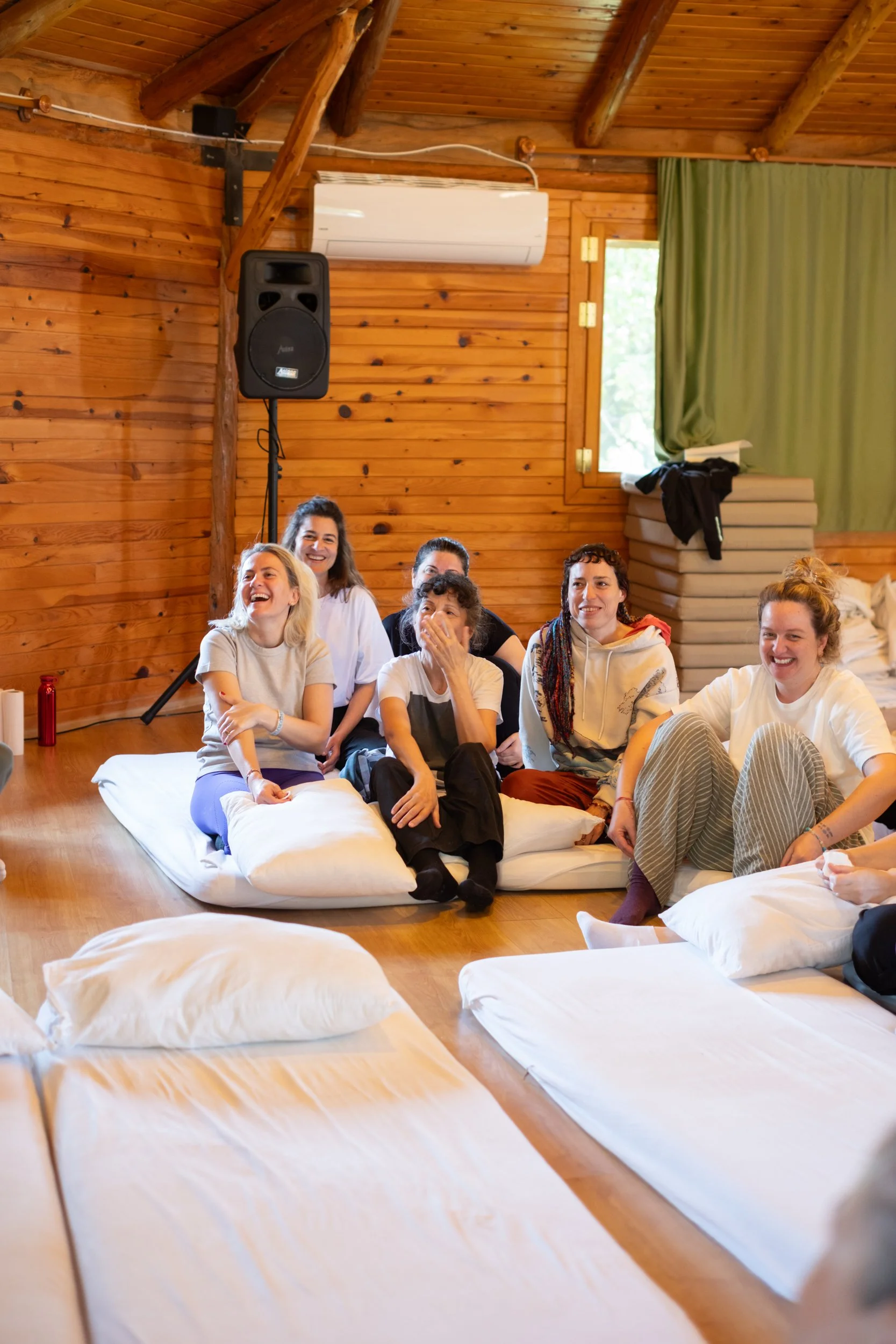 A group of seven women sitting on the floor on white bedding, laughing and smiling in a wooden cabin with green curtains, a window, and a large black speaker. Osho celebration, evening meeting, diamond breath school.