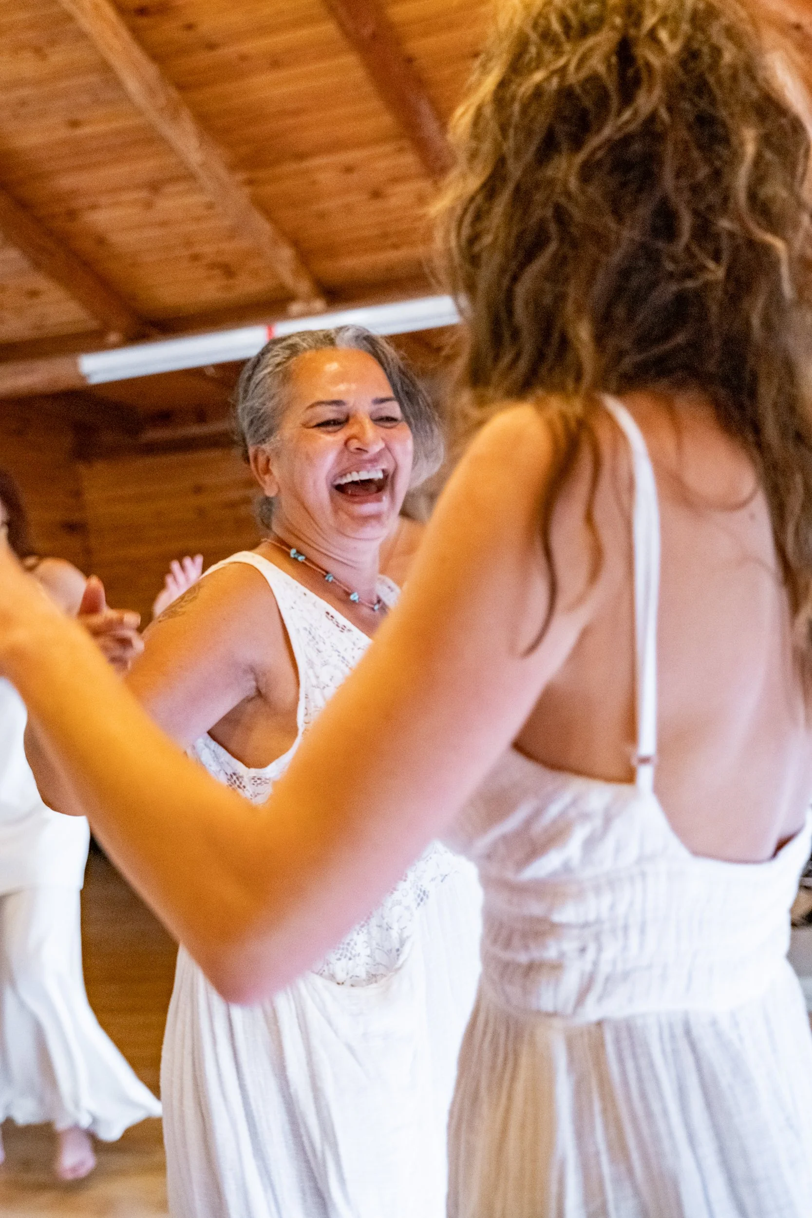 Women in white dresses dancing and laughing inside a wooden room during a celebration or party. Osho celebration, evening meeting, diamond breath school.