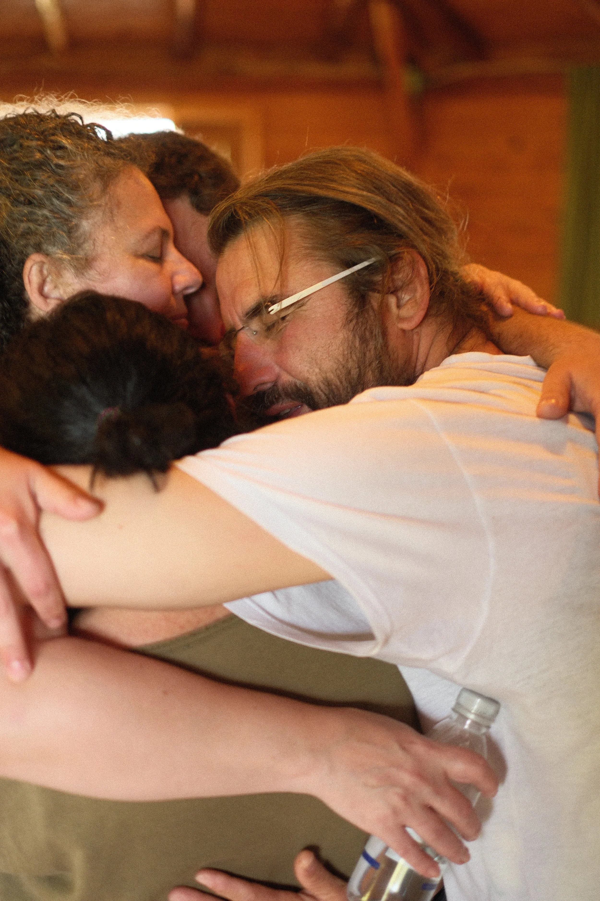 A woman being embraced in a group hug with two others, with a man wearing glasses and a white shirt in the foreground holding a clear medical bottle. Osho celebration, evening meeting, diamond breath school.