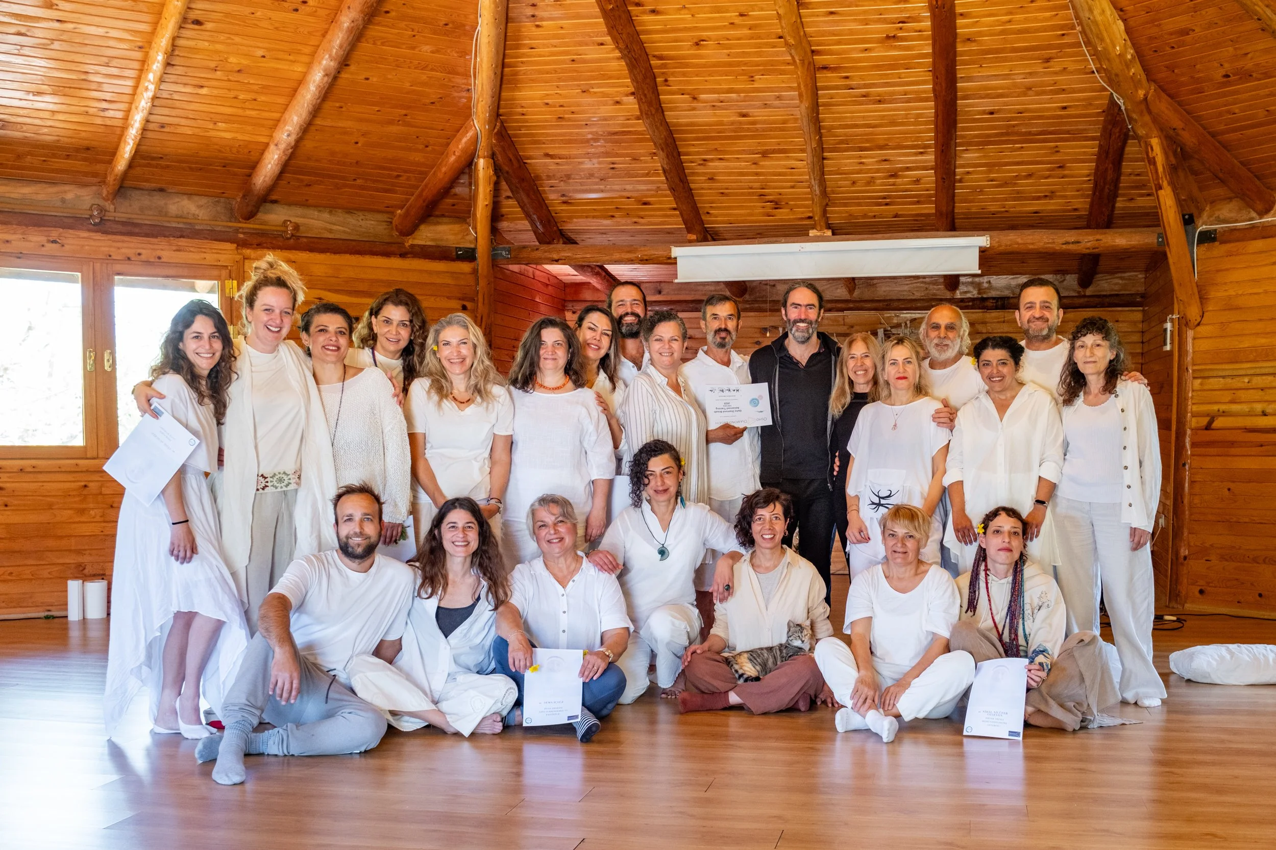 Group of people gathering in a wooden room, smiling at the camera, some holding certificates, dressed in white or light-colored clothing. Osho celebration, evening meeting, diamond breath school.
