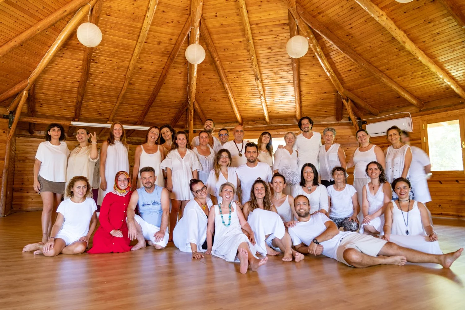 Group of 24 people inside a wooden hall, some sitting and some standing, dressed in white or light-colored clothing, smiling for the photo. Osho celebration, evening meeting, diamond breath school.