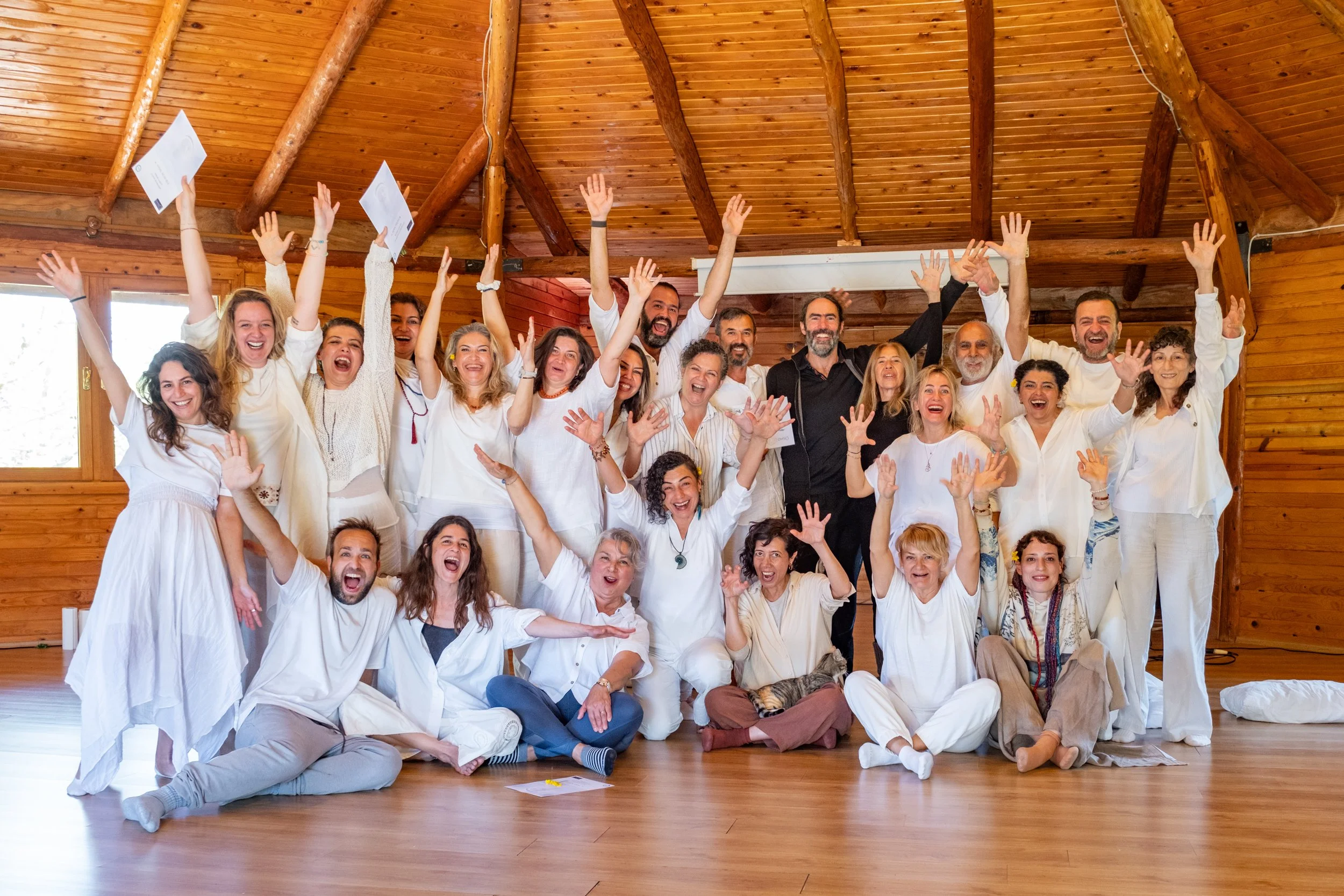 A group of people indoors, celebrating with raised hands, smiling, and some holding papers. The room has a wooden ceiling and walls, with natural light coming through windows. Osho celebration, evening meeting, diamond breath school.