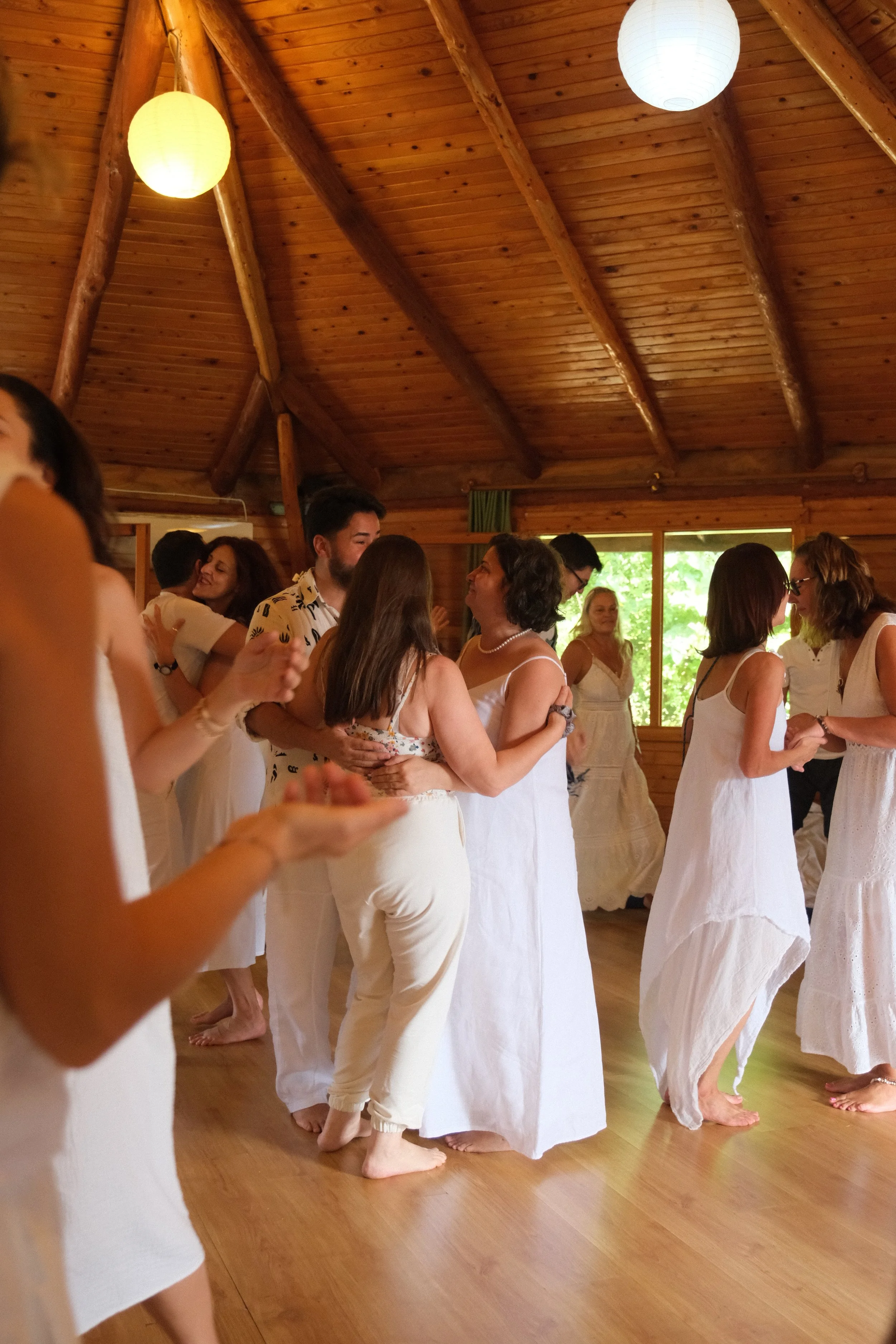 People dancing together in a wooden hall, wearing white dresses, engaged in a celebratory or social dance. Osho celebration, evening meeting, diamond breath school.