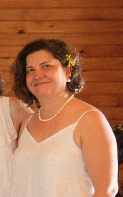 Woman with short curly hair wearing a white dress, pearl necklace, and yellow flower hair accessory, smiling against wooden background.