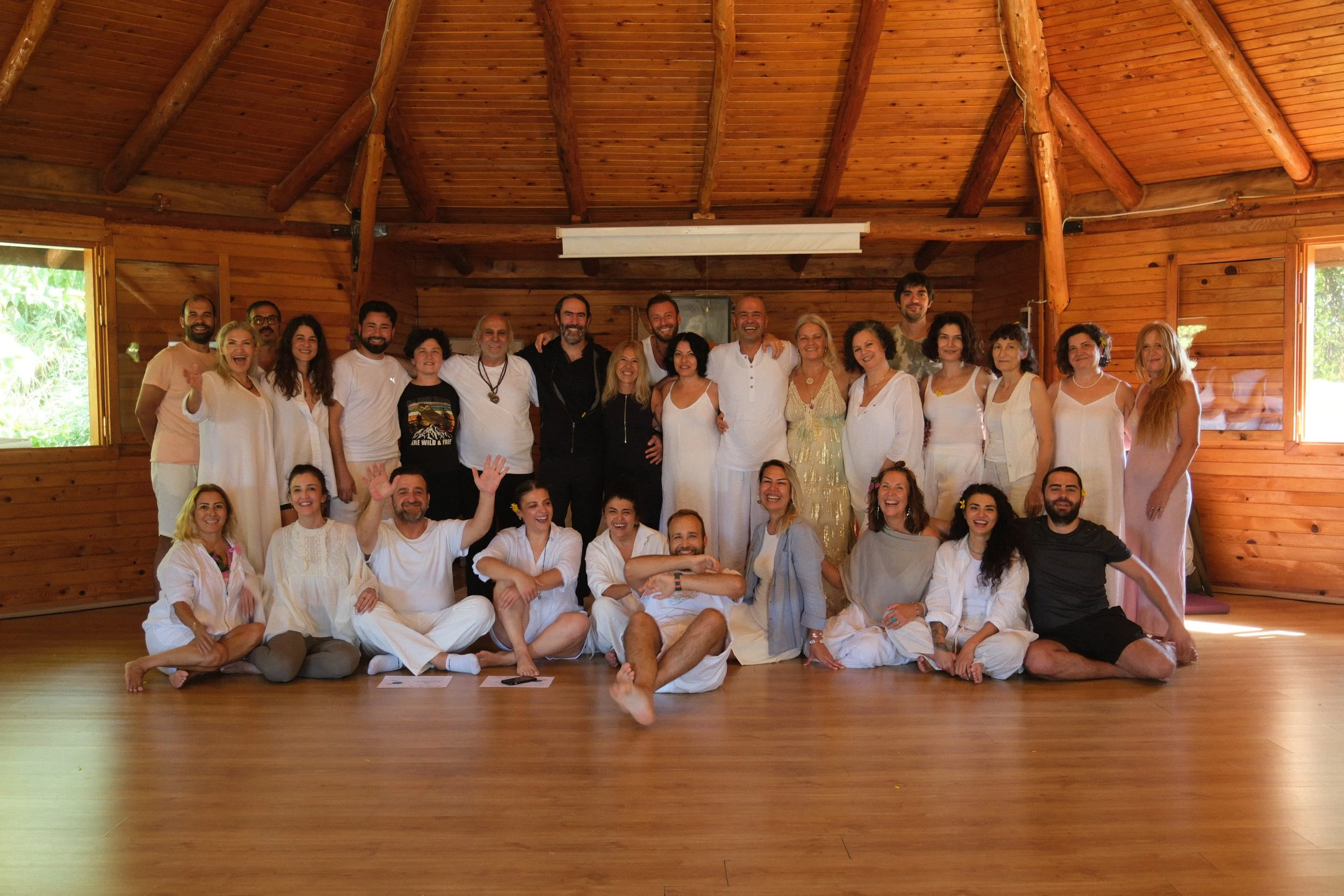 A group of people, some standing and some sitting, gathered in a wooden room with a high ceiling, smiling and enjoying each other's company. Osho celebration, evening meeting, diamond breath school.