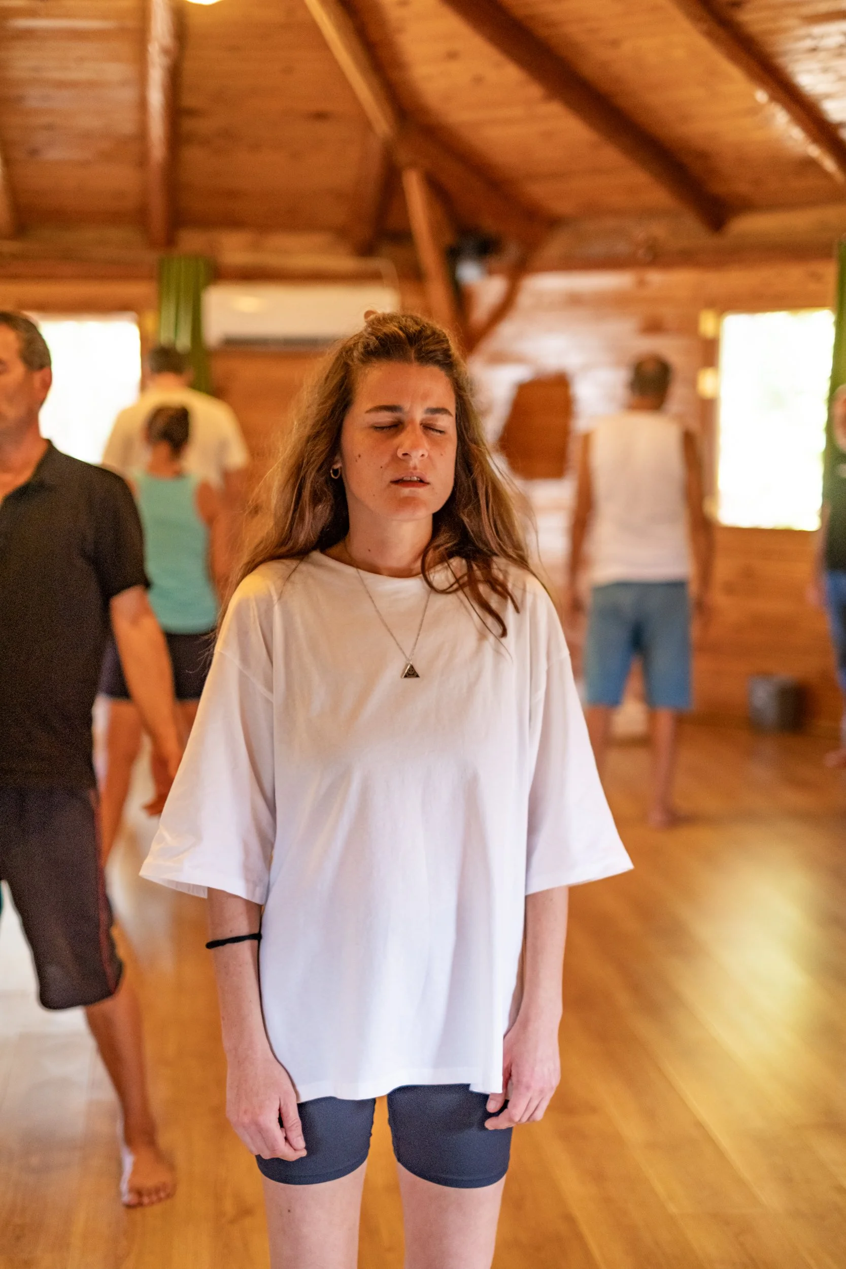 A young woman with long brown hair and wearing a white oversized T-shirt, black shorts, and a necklace, standing in a wooden room with other people in the background, some with closed eyes and others facing different directions.