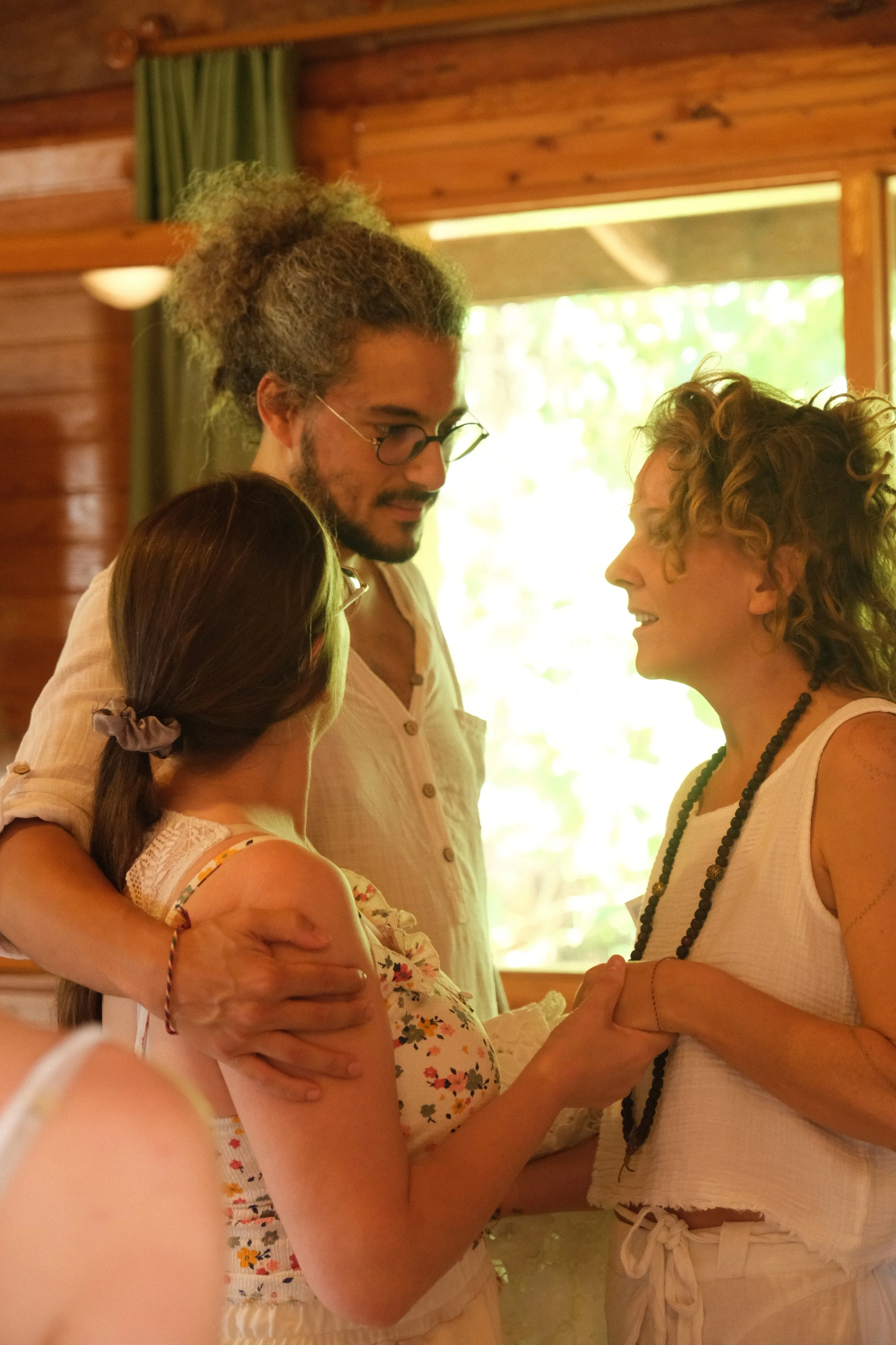 A group of three women standing close together indoors, engaged in conversation and holding hands, with a wooden wall and window with green curtains in the background.