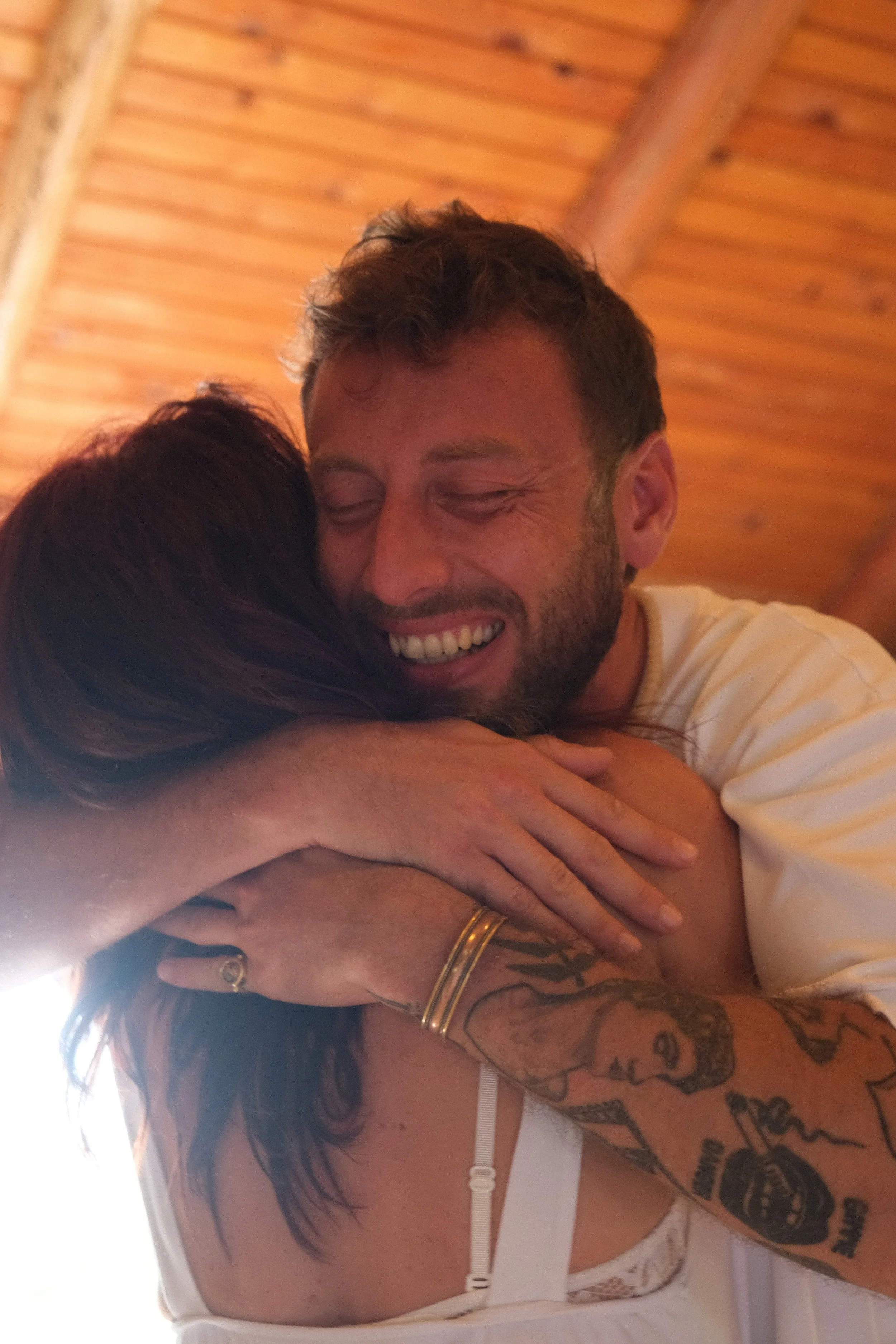 A man with closed eyes smiling, hugging someone, with tattoos on his arm and wearing a white shirt, against a wooden ceiling background. Osho celebration, evening meeting, diamond breath school.