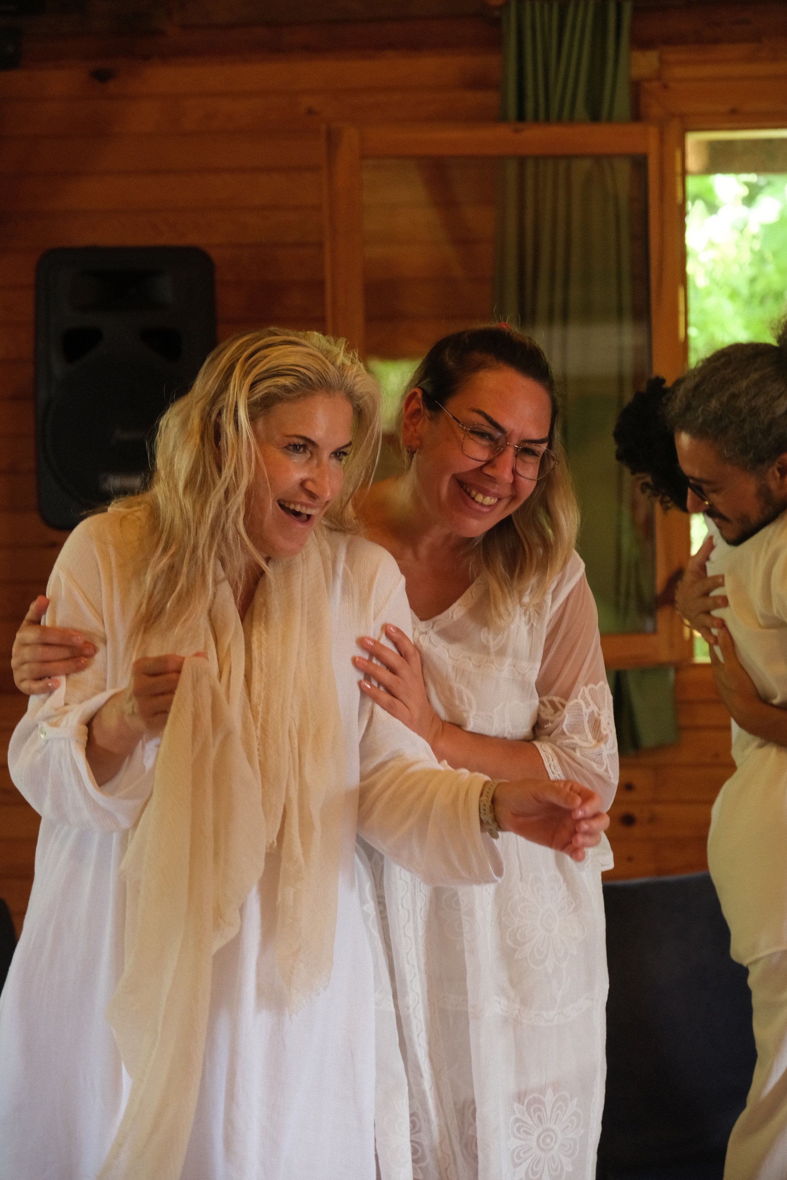 Three women dressed in white, smiling and sharing a joyful moment. Osho celebration, evening meeting, diamond breath school.